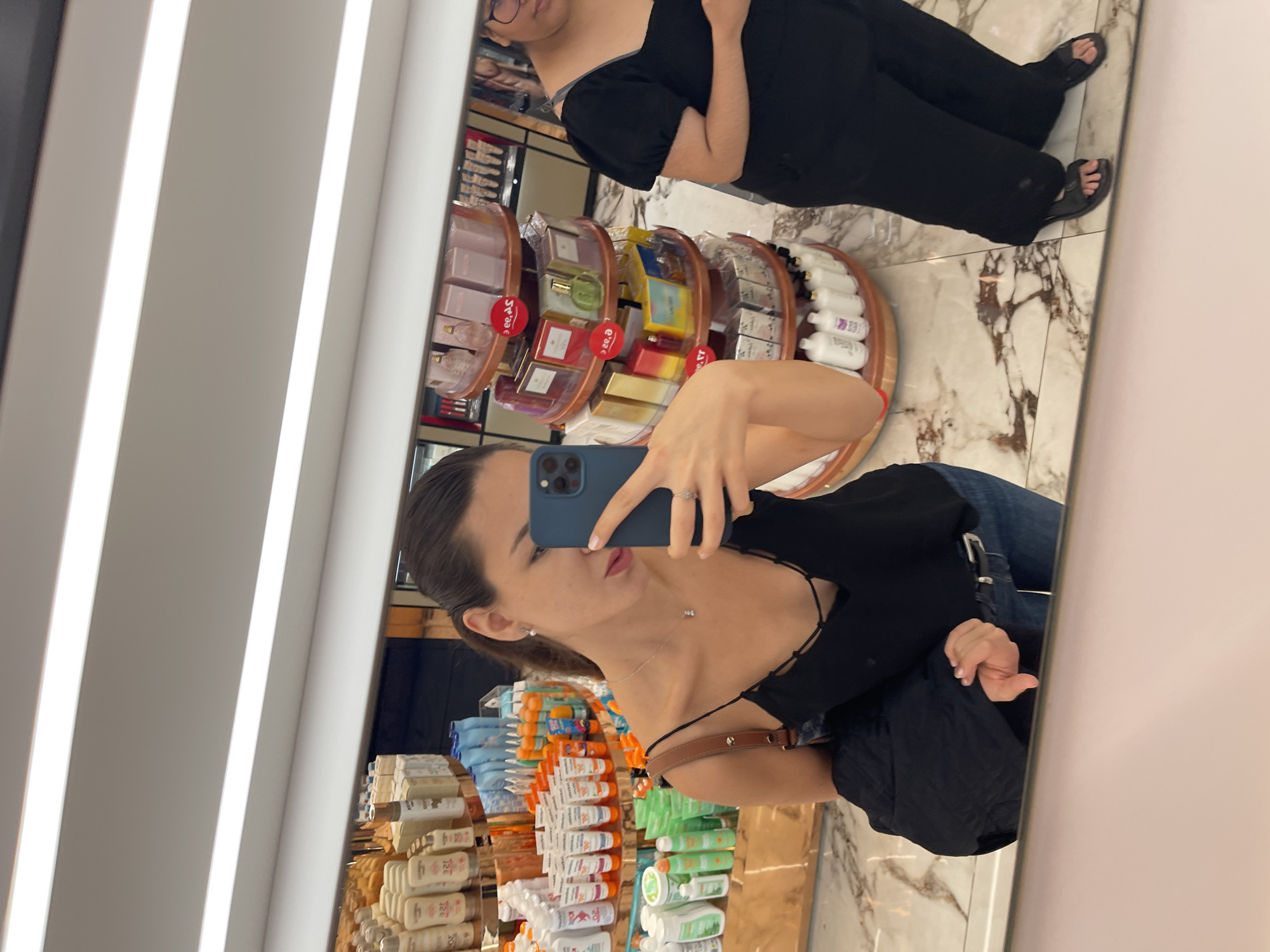 The image depicts a mirror selfie taken inside a retail store, likely a beauty or personal care establishment in Valencia, Spain, given the presence of Spanish text on some product labels. In the foreground, a woman with dark hair pulled back wears a black spaghetti strap top and blue jeans, holding a blue smartphone with her right hand to capture her reflection. She has an earring, a delicate necklace, and a ring visible. A brown shoulder strap is across her left shoulder, and a black garment is draped over her left arm. Her tongue is slightly extended. To her right, a second individual is partially visible, wearing a black puff-sleeve top, black pants, and black sandals, with glasses on. The background, a reflection of the store's interior, features multiple shelves and tiered circular displays stocked with various cosmetics, sunscreens, lotions, and perfumes. The store has white strip lighting on the ceiling and a polished white and grey marble-patterned floor.