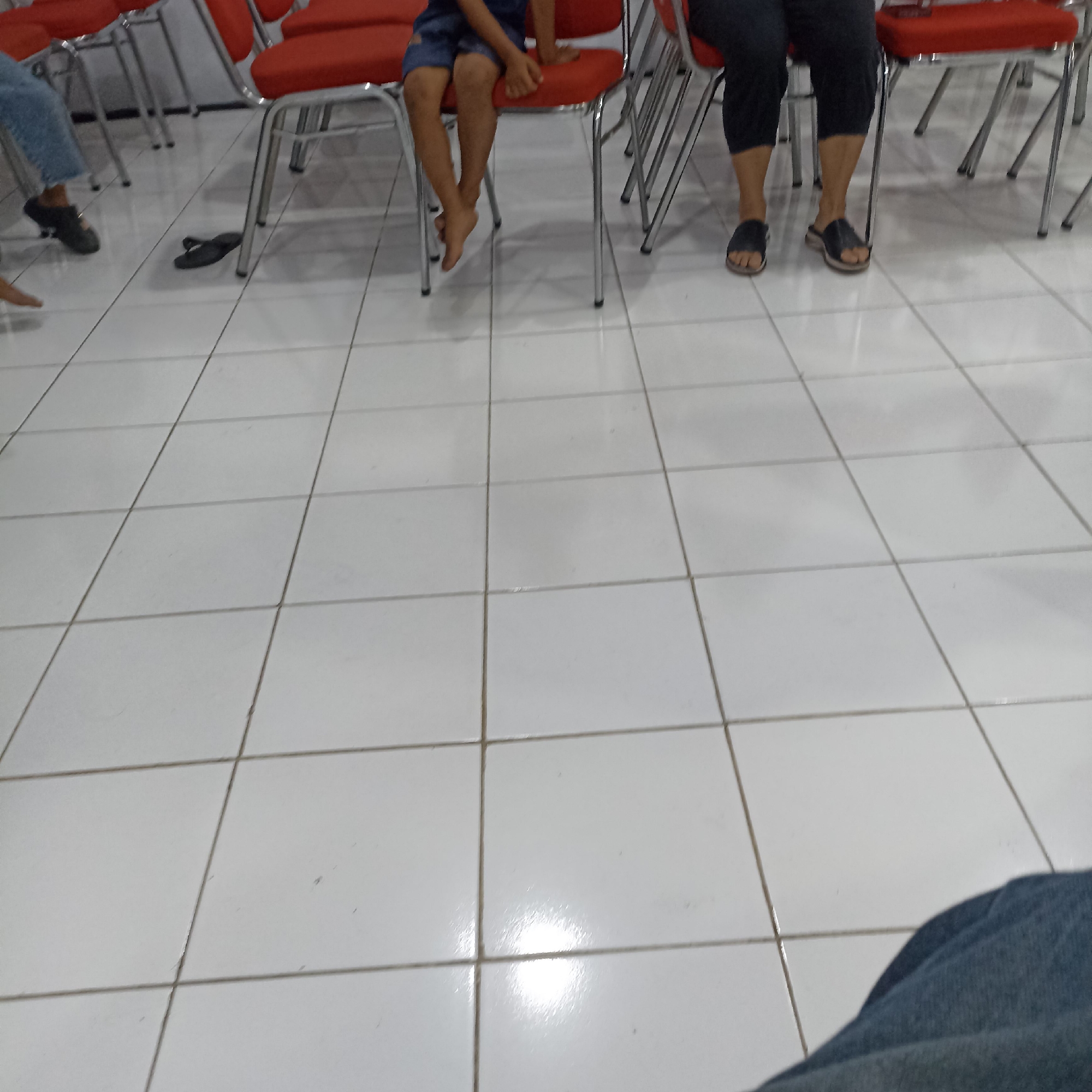 The image depicts a low-angle view of a tiled floor in what appears to be an indoor public space, possibly a waiting area or a hall in Magelang, Indonesia. Several rows of red-upholstered chairs with metal frames are visible in the background. The floor is made of light-colored square tiles with dark grout lines.
In the foreground, the legs of at least three individuals are visible. On the left, a person wearing blue jeans and dark slip-on shoes is partially in frame. To the right of this person, a pair of dark flip-flops are lying on the floor. Further to the right, a child sits on a chair, wearing blue shorts and appears to be barefoot, with their legs crossed and one foot slightly extended. Another adult sits to the right of the child, wearing black pants and black slide sandals. The perspective suggests the photo was taken from a seated or low standing position. The overall lighting is bright, with reflections visible on the tiled floor, indicating artificial lighting or sunlight entering from an unseen source. The context of Magelang, Indonesia, is inferred, but not explicitly visually confirmed within the image itself.