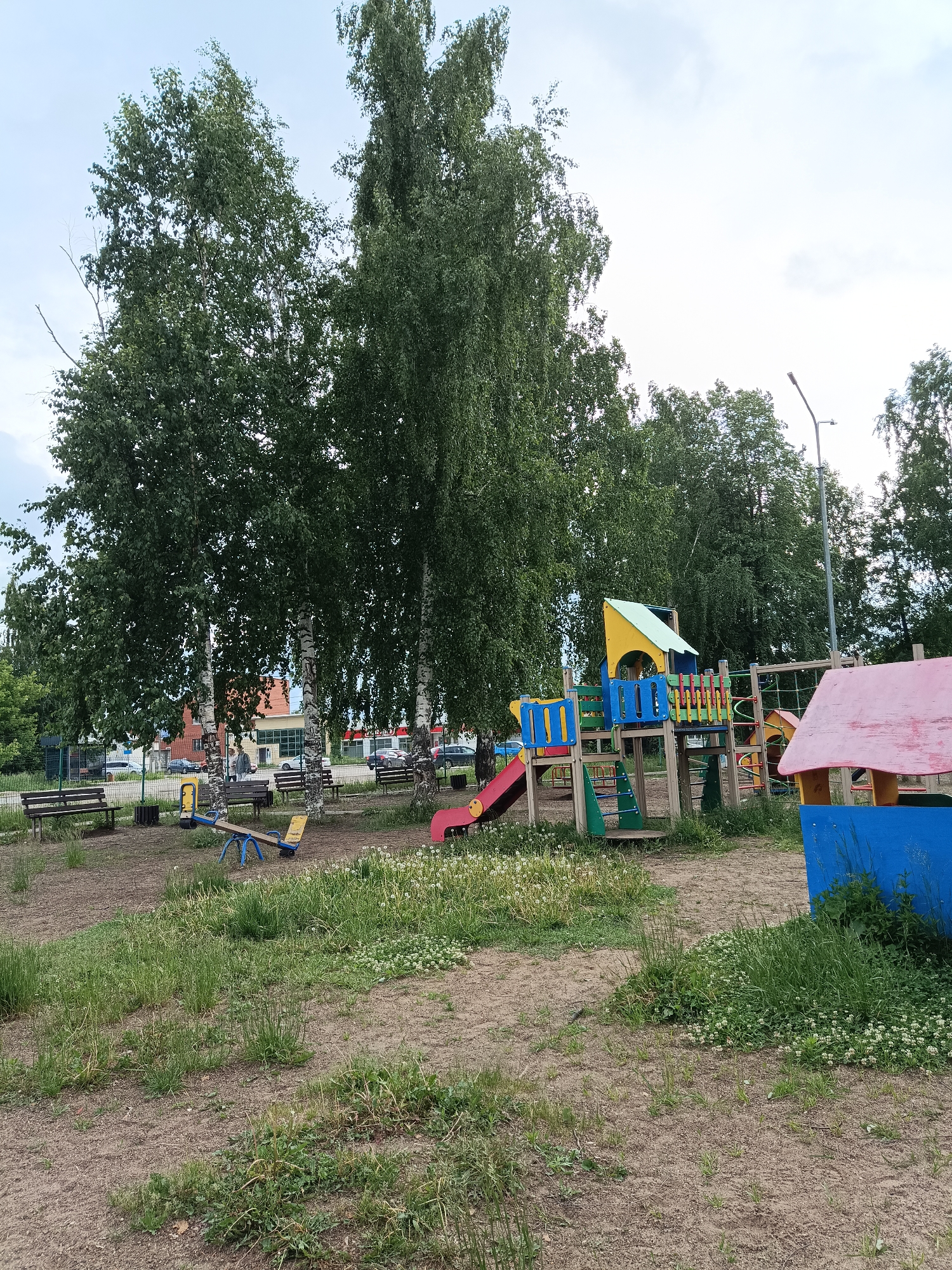 The image depicts an outdoor playground area, likely in Chaikovsky, Russia, given the provided context. The scene is dominated by several tall, leafy trees, some appearing to be birch trees with their characteristic white bark, which cast dappled shade over parts of the area.
In the midground, to the left, there are two wooden benches and a blue and yellow seesaw. Further back on the left, more benches are visible along a fence line, with buildings featuring red roofs and light-colored facades in the distance, suggesting an urban or suburban setting. Several cars are parked or driving in the background.
To the right in the midground, a vibrant children's playground structure stands out with its multi-colored components. It includes a red slide, climbing areas, and a small playhouse with a pink roof and a blue base. A modern street light pole is visible on the right side of the image, extending upwards towards the sky.
The ground covering the playground is a mix of dirt, sparse green grass, and patches of white clover or dandelions, indicating it might be late spring or summer. The sky is overcast with light grey clouds, suggesting either an early morning, late afternoon, or simply a cloudy day.
There are no people present in the image, implying the playground is currently empty and unused. No discernible text is visible on any objects or signs within the frame. The overall impression is that of a quiet, somewhat rustic playground situated within a community.