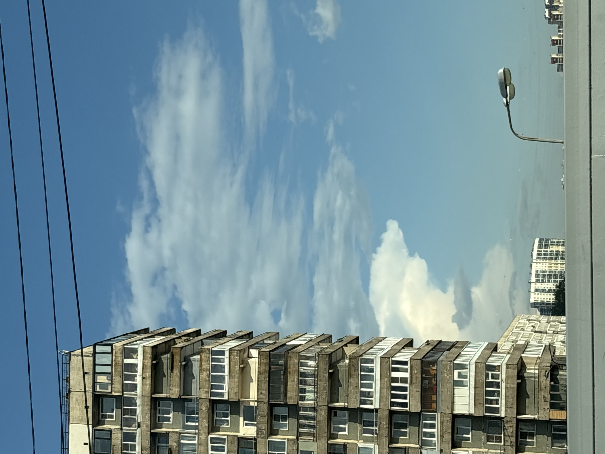 The image captures a view of a high-rise apartment building in Saint Petersburg, Russia, under a bright blue sky with scattered clouds. The building, characterized by its modular concrete structure and numerous balconies with white window frames, dominates the left side of the frame. In the distance, other buildings are visible, including a modern white building with a curved facade and rows of windows. A streetlight with a distinctive dual-head lamp stands on the right, its curved pole extending into the sky. Three dark power lines stretch horizontally across the top of the image. The lower portion of the image shows a light-colored horizontal surface, possibly a barrier or railing. There are no people visible in the scene. The overall impression is one of an urban landscape on a clear day.