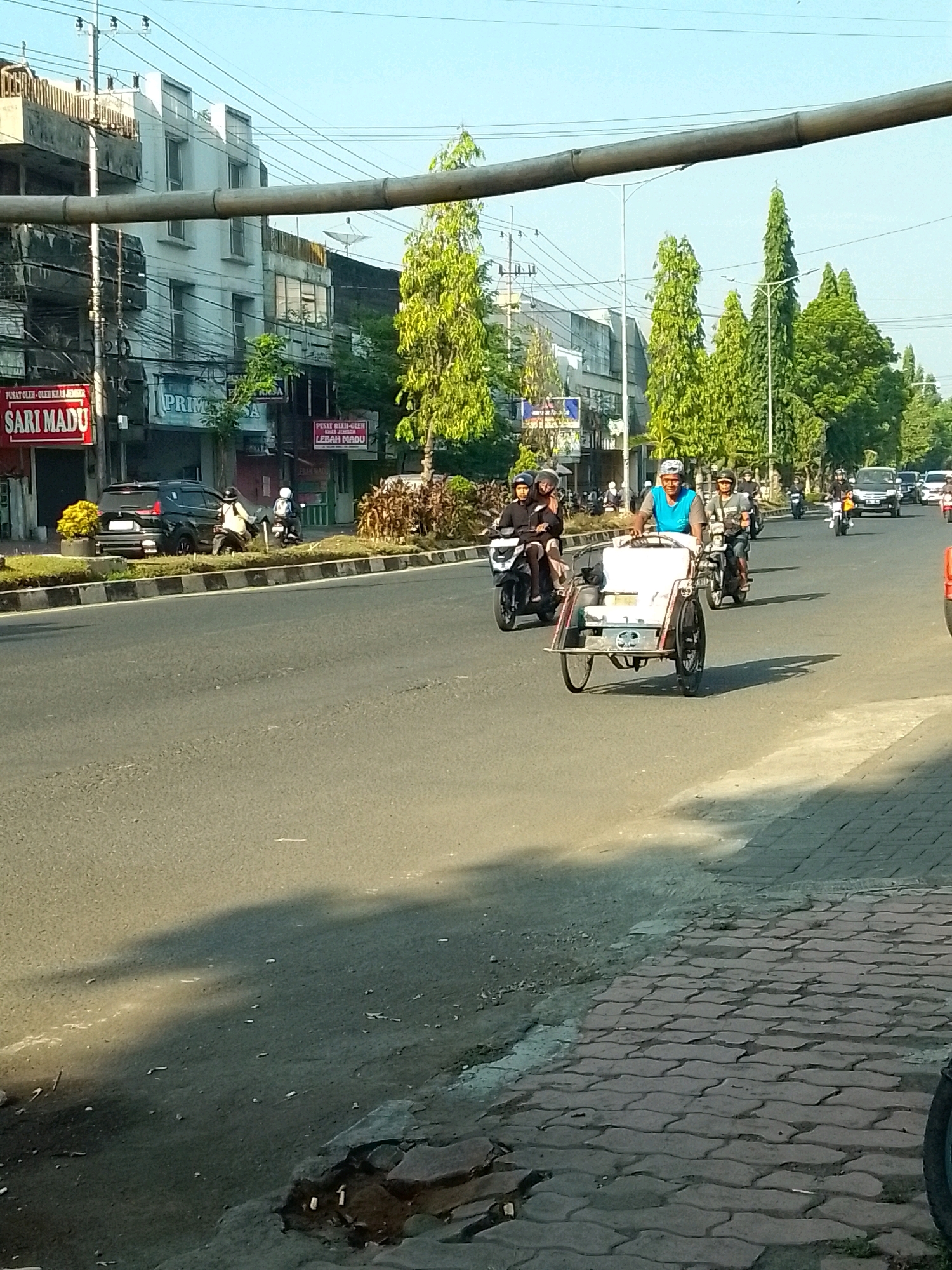 The image depicts a street scene in Jember, Indonesia, under a clear sky. In the foreground, a brick sidewalk runs along the right side of the frame. The street is paved and features multiple lanes of traffic. Several vehicles are present, including motorcycles, cars, and a pedicab. On the left side of the street, buildings with storefronts are visible, one of which has a red sign with white text that reads "SARI MADU". The buildings are multi-story with visible windows and electrical wires running overhead. Green trees line parts of the street, and landscaping with grass and shrubs is also present.
In the center of the image, a pedicab is being ridden by a man wearing a blue shirt and a cap. He is moving from left to right across the frame. Several motorcycles are also in motion. One motorcycle in the middle distance has two riders, both wearing helmets. Another motorcycle is approaching from the distance with a single rider. A black car is parked on the side of the road near the buildings. Shadows from the overhead structure and trees are cast across the road. The overall impression is a bustling street during daylight hours.