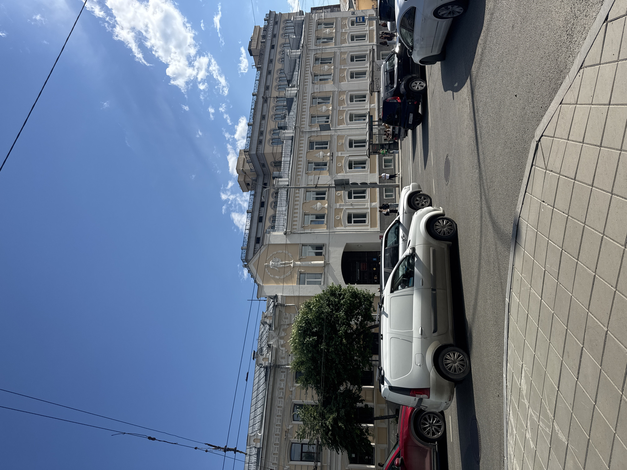 The image depicts a sunny day in Yekaterinburg, Russia. A tall, ornate building with cream-colored facade and numerous windows stands on the left side of the frame. The building's architecture suggests it is historical. Trees with lush green foliage are visible in front of the building. A street runs alongside the building, with several cars parked along the curb. These include a white van, a black SUV, and a red car. The sidewalk is paved with square tiles. Overhead, telephone or power lines crisscross the bright blue sky, which is dotted with fluffy white clouds. The scene appears to be a quiet street in a city, with no specific activity or event clearly taking place, beyond the presence of parked cars and a few indistinct figures on the sidewalk. The lighting indicates it is daytime, likely mid-morning or afternoon, given the strong sunlight and clear skies. Text is visible on the building, reading "PAROVSKY".