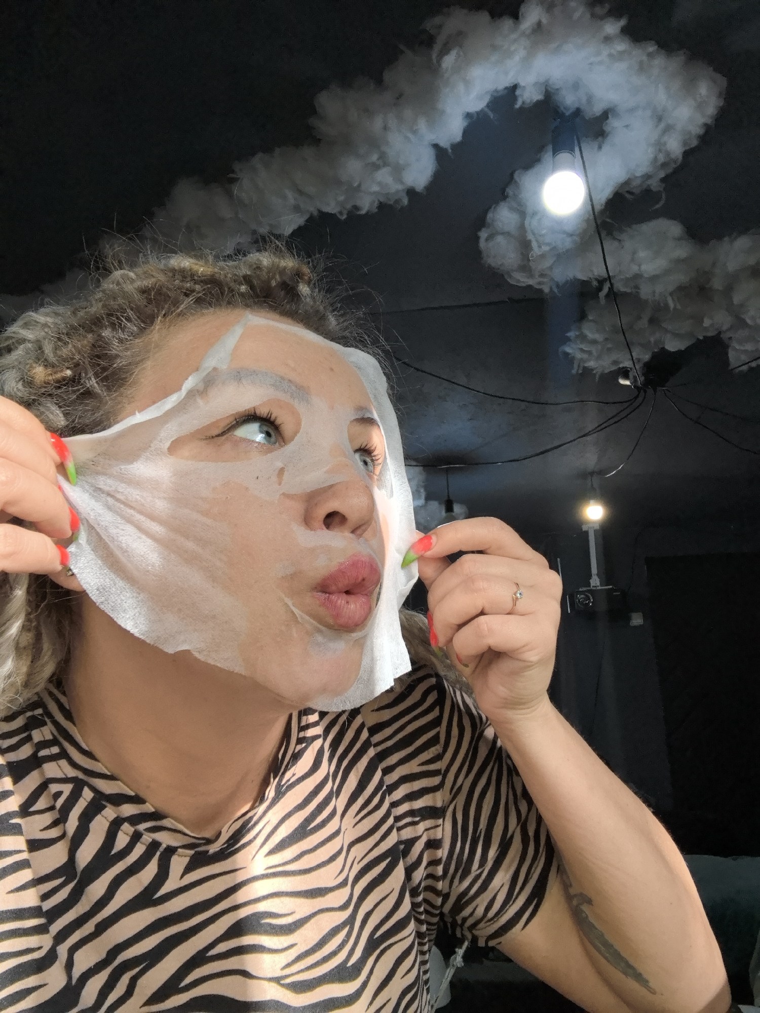A woman with curly blonde hair and a facial mask is peeling it off her face. She has green and red nails. She is wearing a tan and black zebra print shirt. The background is a black ceiling with white cloud-like decorations and exposed light bulbs with wires. The location is not evident and there is no indication it is the city of Noviy, Russia.