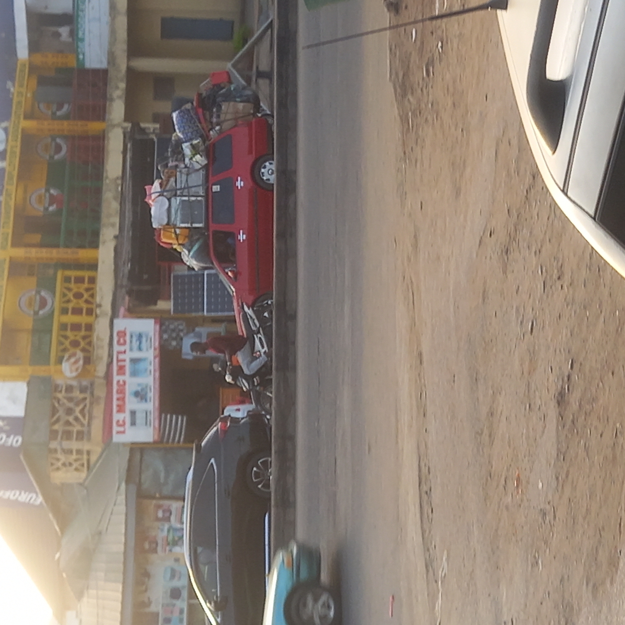 The image shows a street scene in what appears to be Sardauna, Nigeria. A red vehicle is heavily loaded with goods on its roof. A motorcyclist is seen driving on the road next to the red vehicle. Parked nearby are a dark-colored SUV and a light blue car. The roadside has a building with signage that reads "LC. MARC INTL CO." There are people present, including the driver of the motorcycle and possibly occupants of the red car. The setting appears to be a commercial area with buildings and vehicles on a street.