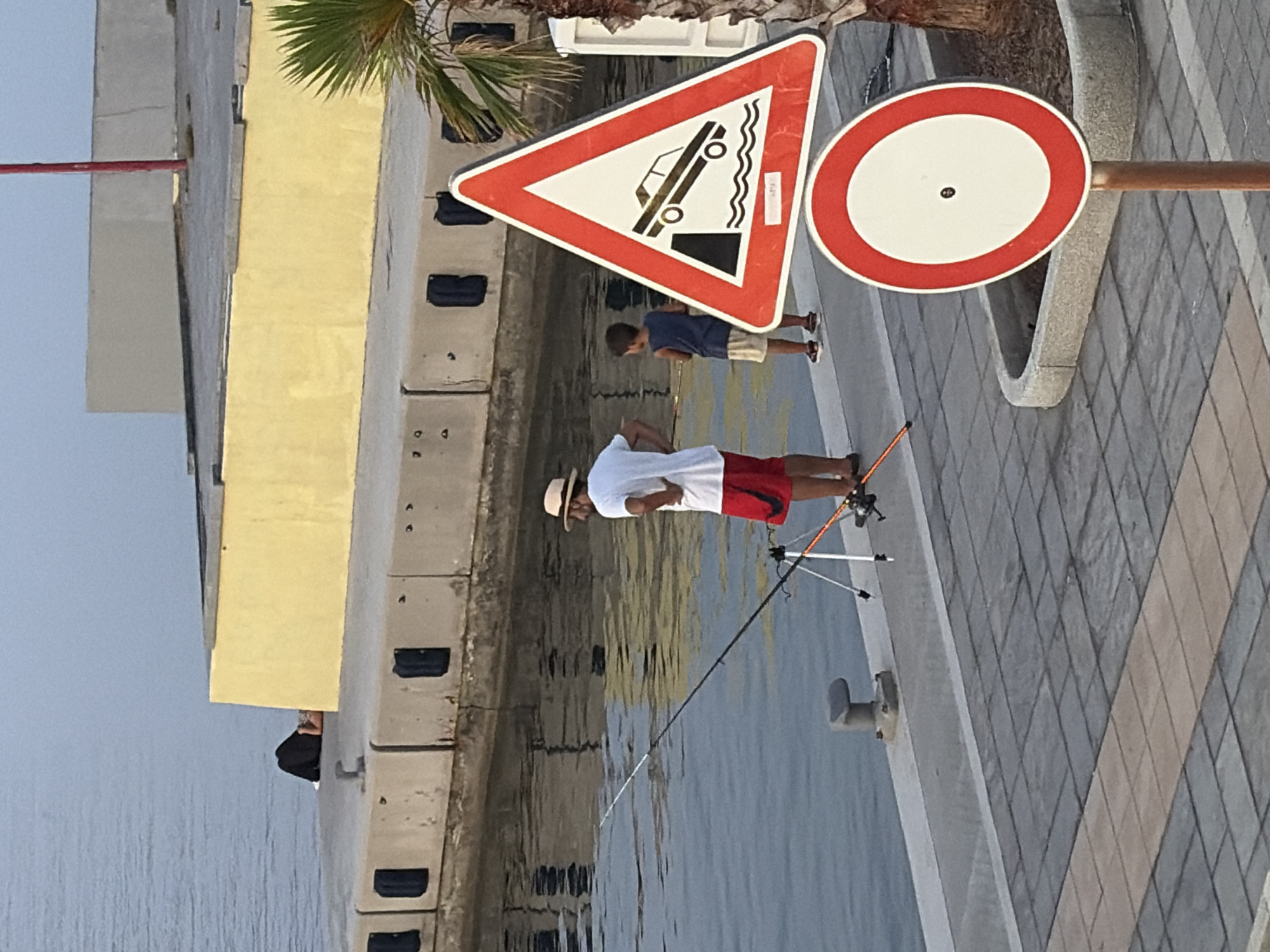 This media file depicts a waterfront scene in Imperia, Italy. The primary subjects are two people: a man fishing and a child. The man, wearing a hat, white shirt, and red shorts, is actively fishing with a rod and reel set up on a tripod. The child, in a blue shirt and light shorts, stands behind him, observing.
The setting is a paved walkway next to the water, likely a harbor or marina. A yellow building is visible in the background, along with some concrete structures. There are two prominent traffic signs. The first is a triangular warning sign with a red border and a white interior, displaying a black pictogram of a car tilting into the water, indicating a drop-off or submerged edge. The second is a circular prohibition sign, also with a red border and a white interior, featuring a black dot in the center, which typically signifies "no stopping" or "no entry" depending on context and accompanying signs.
The lighting suggests it is daytime, possibly late afternoon, with the sun casting shadows. The water appears calm, reflecting the surrounding structures and sky. The overall atmosphere seems peaceful, with the focus on the activity of fishing. There is no visible text on the clothing or other elements in the image.