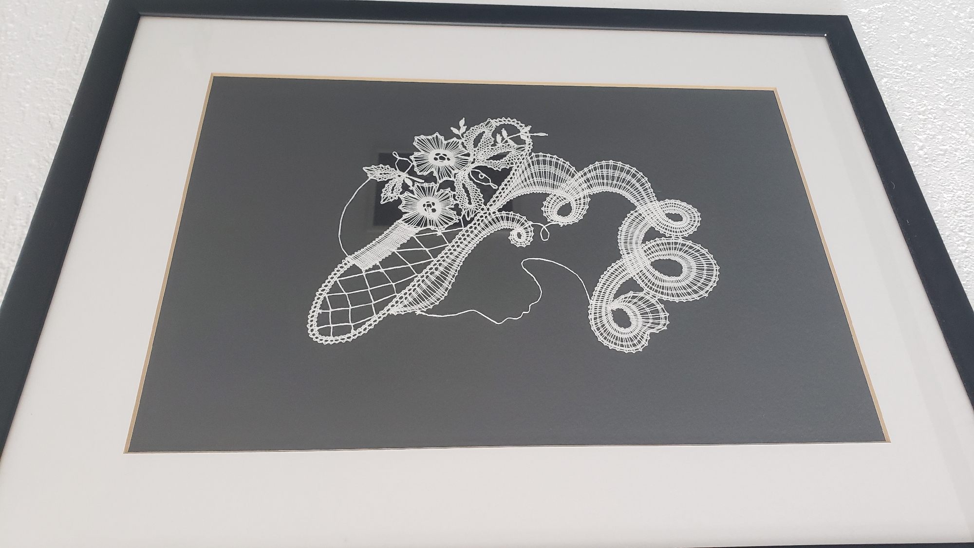 The image displays a framed piece of art featuring a white lace silhouette of a woman's profile, adorned with a decorative hat and flowing hair. The artwork is mounted on a dark gray background and framed with a wide white mat and a black outer frame. The background wall is textured and white. The artwork is hanging on a wall in a room, likely a home or an art gallery. There is no discernible information to indicate the specific location within the city of Prešov, Slovakia.
