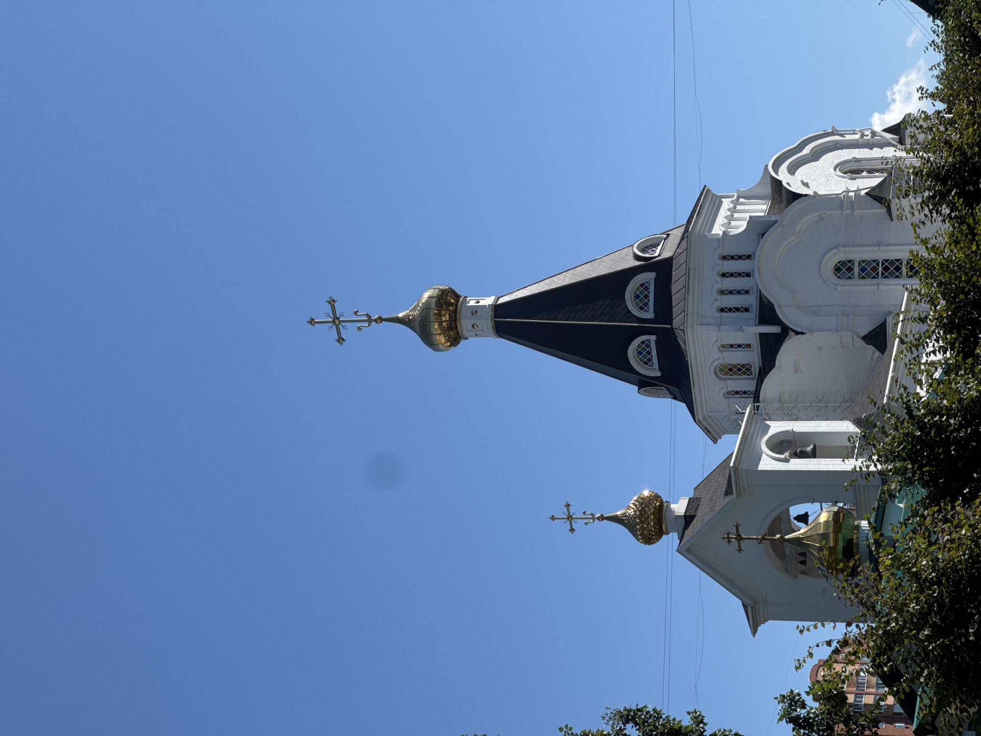 An exterior shot shows a white church with a black roof and golden domes under a clear blue sky. The church has multiple architectural elements, including arched windows with stained glass, a bell tower with bells, and several onion domes topped with crosses. Lush green trees are in the foreground, partially obscuring the lower portion of the church. In the background, a multi-story brick building is visible, suggesting an urban or suburban setting. The image captures the ornate details of the church's exterior against a serene sky.