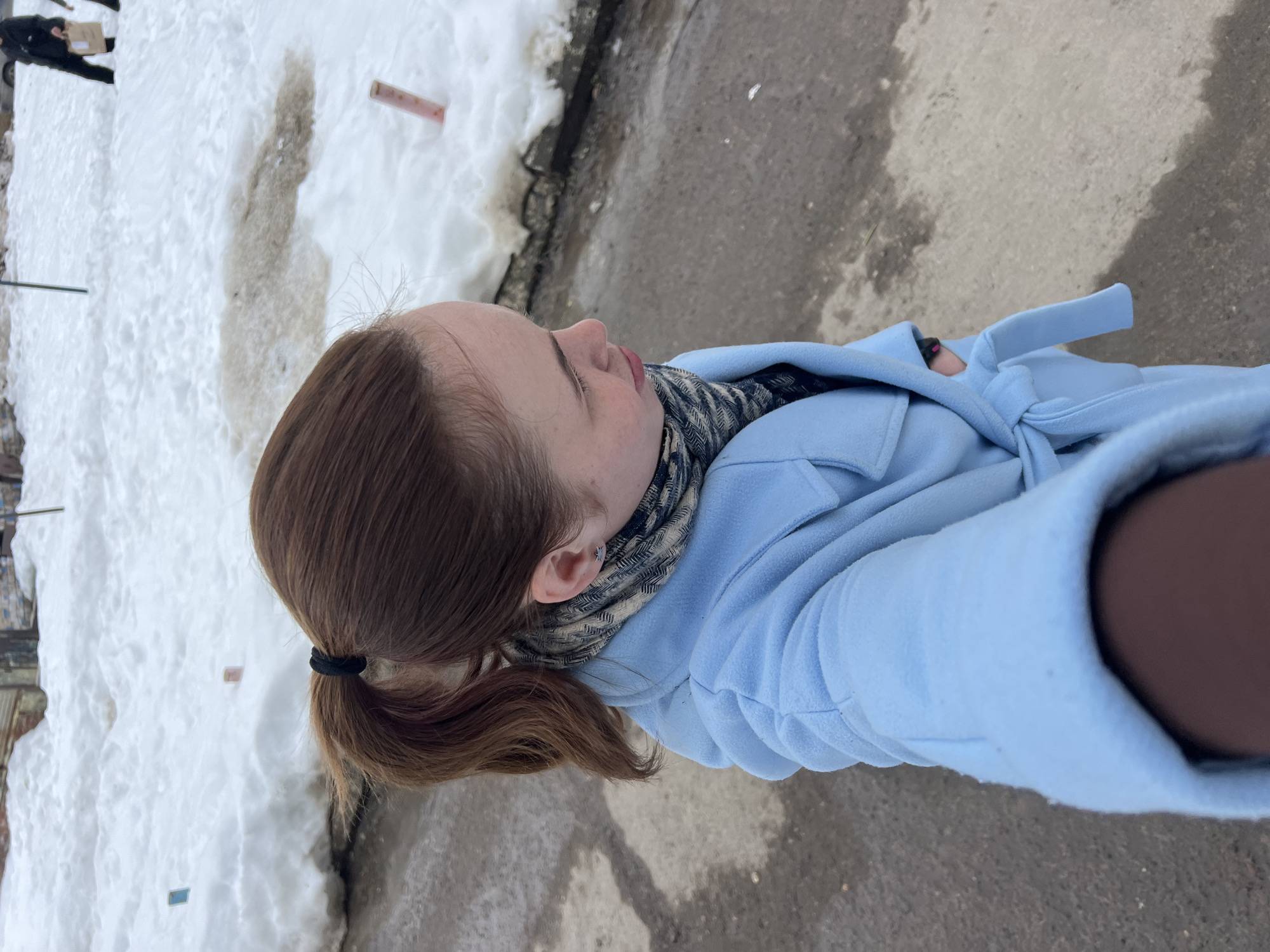 A medium shot captures a young Caucasian woman with light-brown hair pulled back in a ponytail, standing outside. She wears a light blue coat tied at the waist and a scarf around her neck. Her head is turned towards the left, and she appears to be holding the camera herself, possibly taking a selfie. The ground around her is a mix of concrete and patches of snow. In the background, there is more snow along with some urban structures. It is daytime. The location is identified as Chaikovsky, Russia.