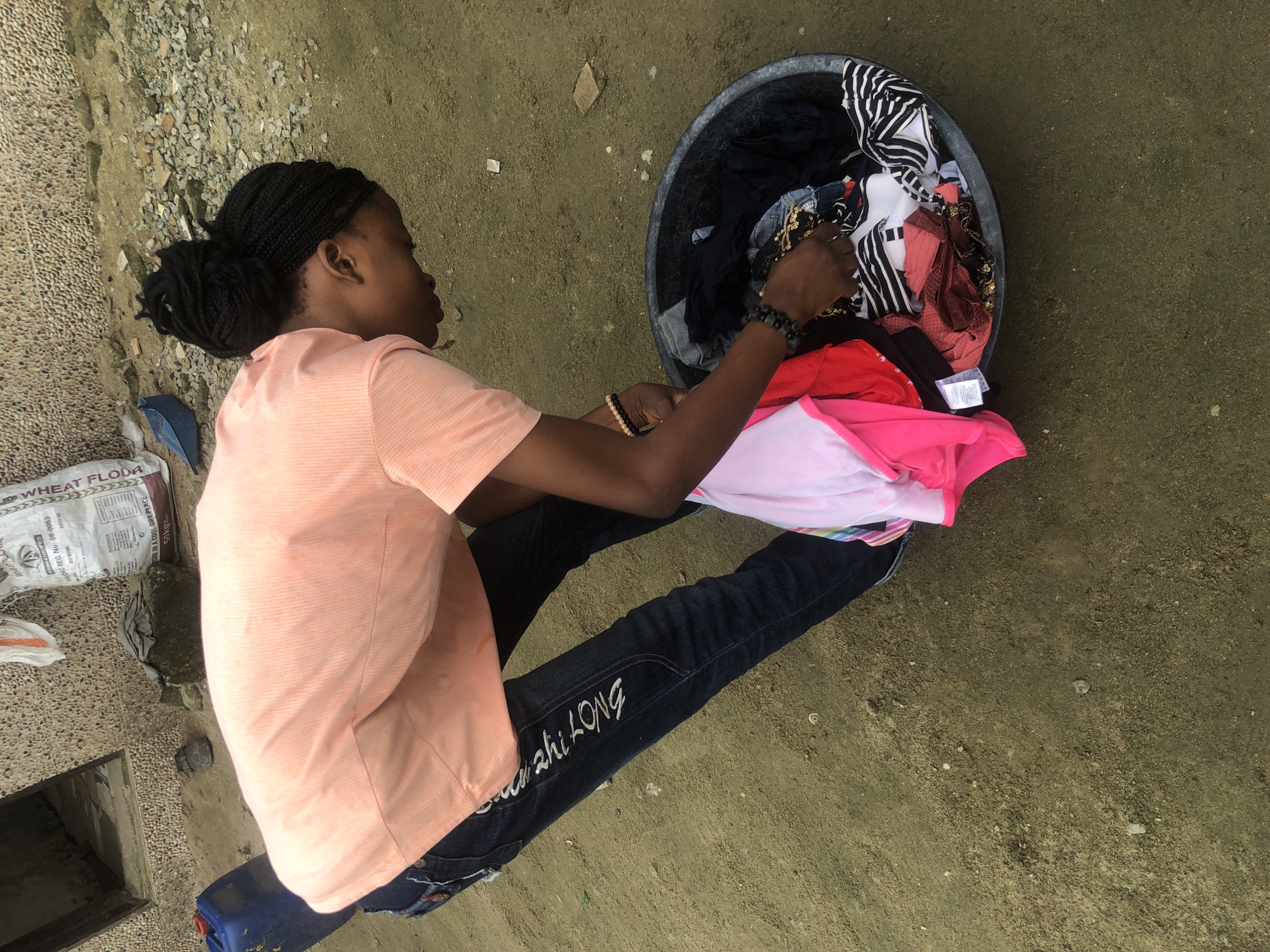 An individual is shown in Ado, Nigeria, bending over a large basin filled with laundry. The person wears a light peach t-shirt and dark blue jeans with white embroidery on the left leg. They are interacting with the contents of the basin, which includes various items of clothing such as pink, white, and red tops, as well as striped and dark-colored garments. The ground appears to be a mixture of dirt and gravel. In the background, there is a building with a textured stone facade, a doorway, and a large white bag of what appears to be wheat flour. A blue plastic container is also visible to the left of the person. The overall scene suggests an outdoor domestic chore taking place in a somewhat unkempt environment.