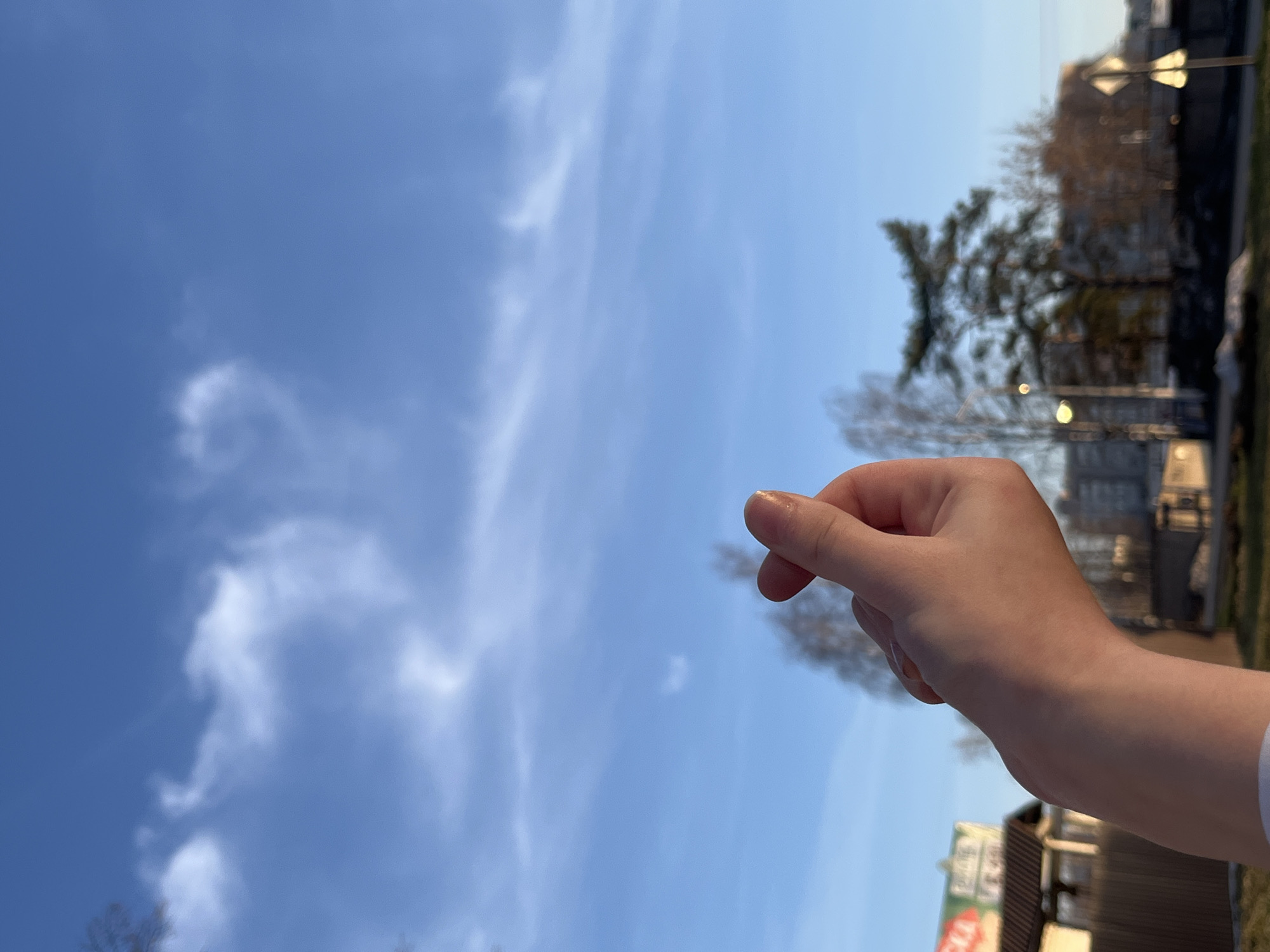 A hand is positioned in the foreground against a bright blue sky with wispy white clouds. The hand is fair-skinned, with the index finger and thumb pinched together. In the background, slightly blurred, are trees, buildings, and what appears to be a streetlight. A sign with illegible text is also visible. It is not possible to confirm the location with the information given.