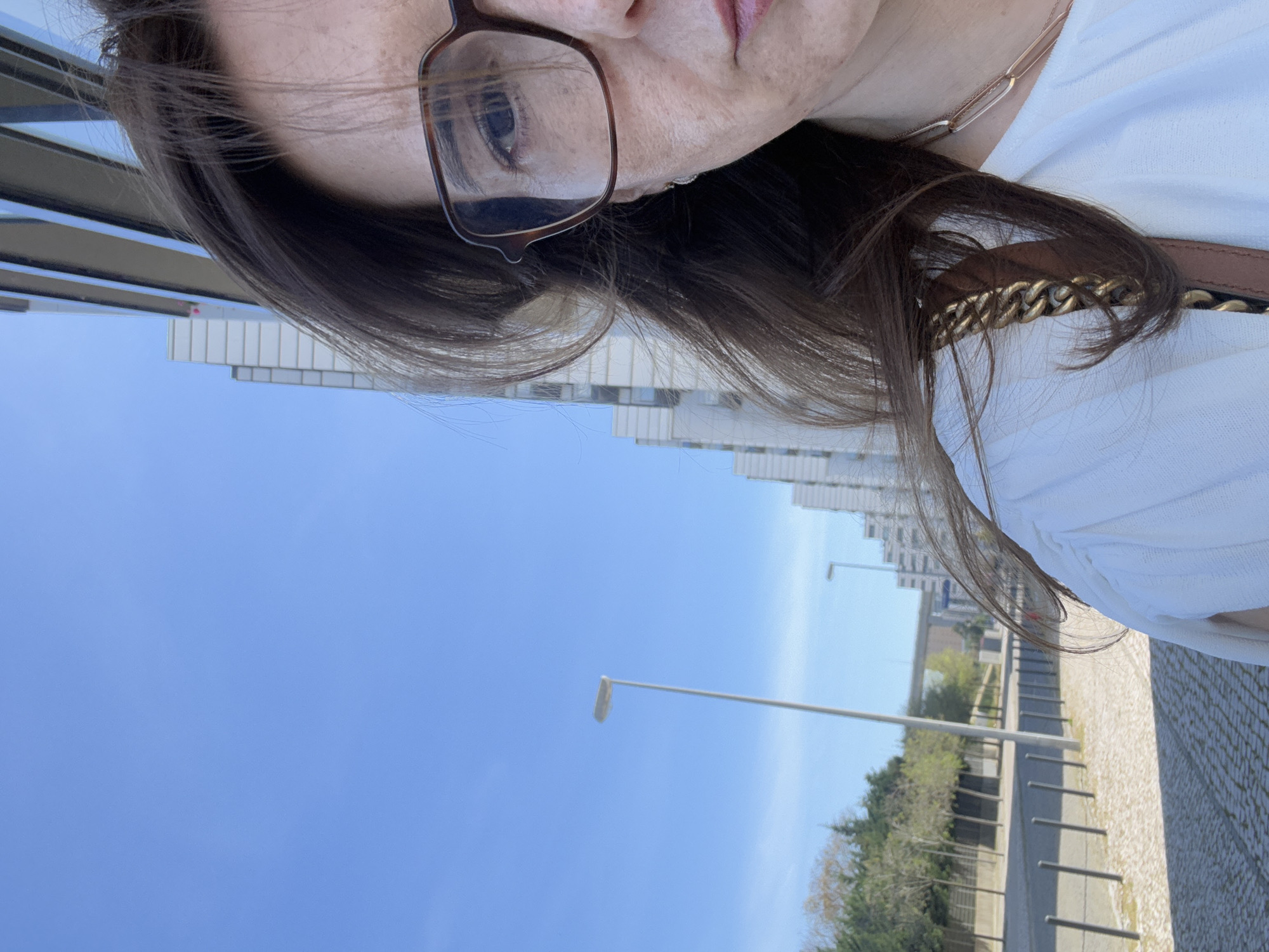 A woman wearing glasses and a white shirt is taking a selfie. The background shows a street with trees, a sidewalk paved with grey bricks, and modern buildings. A lamppost is visible against a clear blue sky. The location is likely Odivelas, Portugal, based on the urban landscape and architectural style.