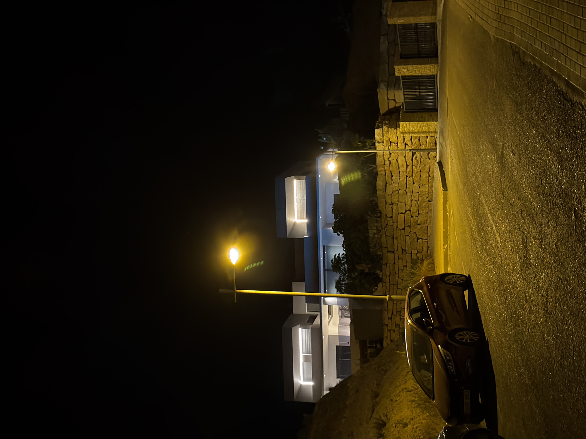 A dark, wide-angle, night photograph captures a street scene in Finestrat, Spain. The image is dominated by artificial light cast from a streetlamp in the mid-ground and illumination from a modern, multi-story house in the background. The house, with its minimalist architectural style, features prominent horizontal lines and illuminated windows, suggesting a contemporary design. In the foreground, a dark red hatchback car is parked on the asphalt road. To the left of the car, another vehicle is partially visible. The street is bordered by a low, rough stone wall along its right side, behind which a section of the property is visible, including large metal gates. The terrain to the left of the street slopes upwards, covered in sparse vegetation and rocks, characteristic of the Mediterranean landscape. The overall atmosphere is one of quiet solitude under the artificial glow of the night.