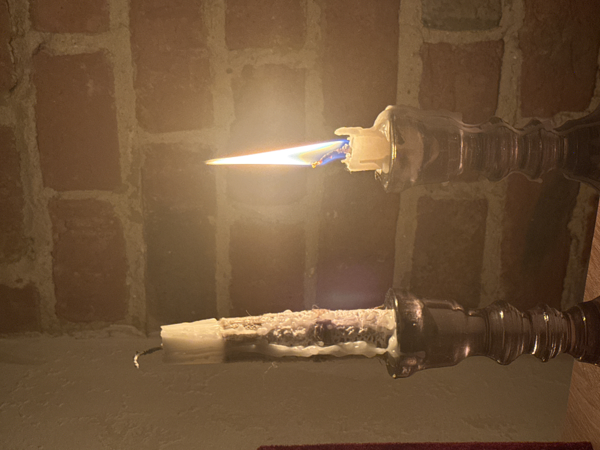 This image captures an intimate indoor scene featuring two candles in glass candlesticks against a textured background. The top candle is actively burning, emitting a bright, elongated flame with distinct blue hues near the wick, indicating combustion. The candle itself is white, partially melted, and housed in a clear, ribbed glass holder.
Below it, a second, unlit white candle rests in a similar, clear glass candlestick. This lower candle is significantly encrusted with solidified, melted wax, suggesting it has burned for a considerable time in the past. Its wick is dark and appears to be extinguished or unused.
The background consists of a reddish-brown brick wall with lighter mortar lines, suggesting an exposed brick interior, possibly part of a fireplace or an architectural feature. Beneath the lower candle, a lighter-colored, smooth surface, potentially a mantelpiece or shelf, is visible. A sliver of dark red fabric is just perceptible at the very bottom edge of the frame.
The overall lighting is dim, with the sole source of illumination being the flame of the upper candle, casting a warm, soft glow across the scene. The image evokes a calm and cozy atmosphere, likely captured in the evening or at night. There is no discernible text in the image. The scene is set in Rostov-on-Don, Russia.