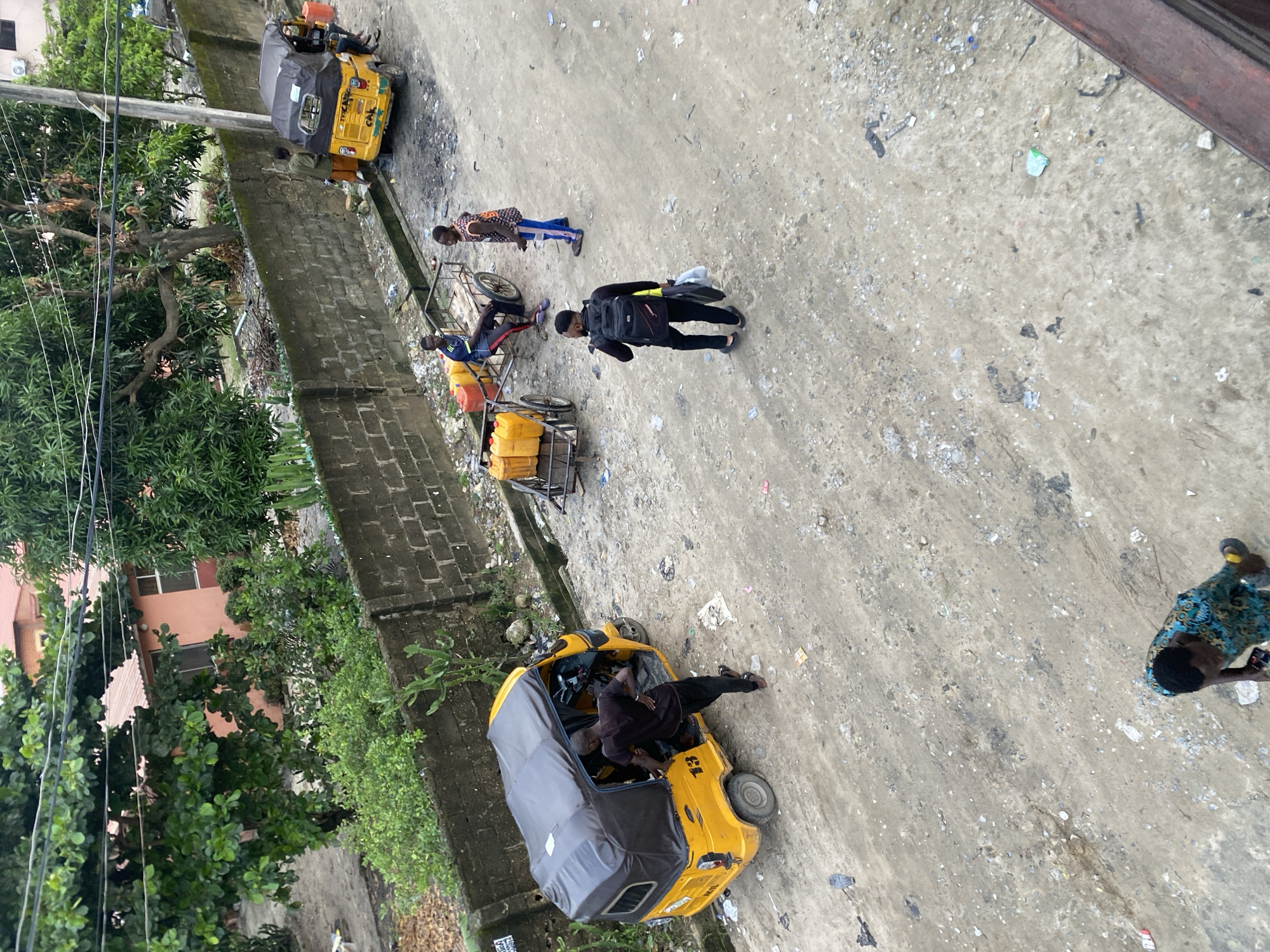 Captured from an elevated perspective in Ado, Nigeria, the scene features a street where a yellow three-wheeled vehicle is parked. Adjacent to the vehicle, a person is visible inside and another leaning against it. Further up the street, two people stand beside a cart filled with yellow containers. A person wearing black clothing and carrying a backpack walks past them. Additionally, another three-wheeled vehicle is positioned at the top of the street. The street is littered with debris and waste. Green foliage and buildings frame the left side of the image.