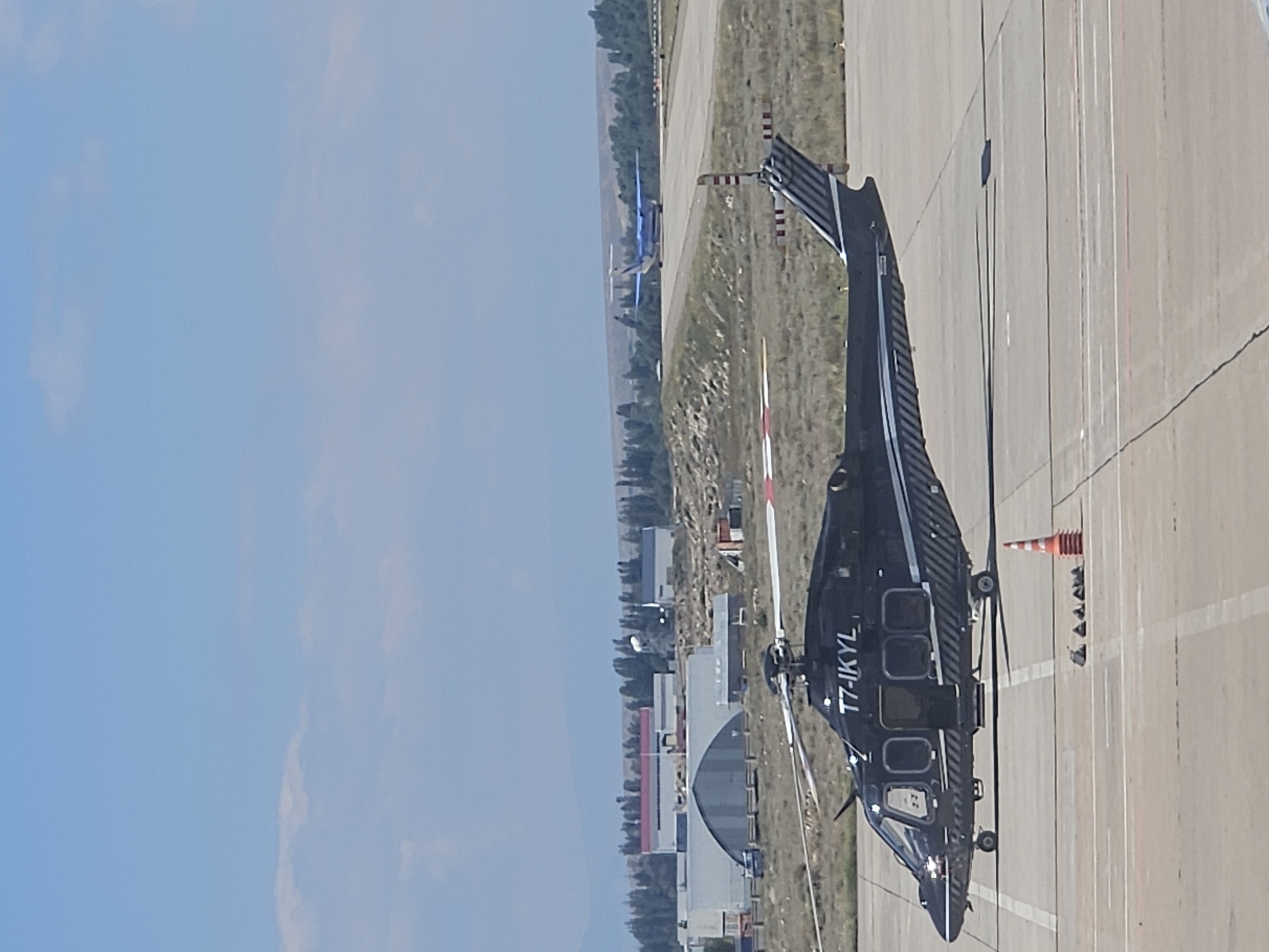 A black helicopter with the registration T7-IKYL is parked on an asphalt tarmac. The helicopter is dark in color with white stripes along its side. Several windows are visible on the side of the helicopter, and the main rotor is extended. In the background, a blue airplane is on a runway. Further in the distance, there are buildings and trees. The sky is clear and blue with a few scattered clouds. The scene appears to be an airport or aviation facility.