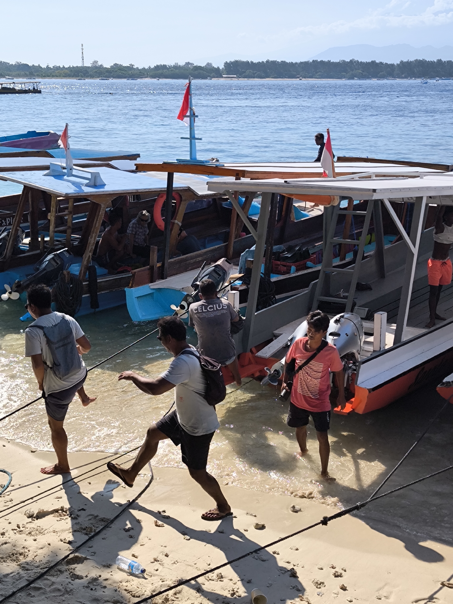 A medium-shot captures several people walking along a sandy beach next to a row of traditional Indonesian boats in an unknown city of Indonesia. The boats are wooden with blue and gray accents, some with red and white Indonesian flags. People are boarding and disembarking the boats. The water is clear and blue, with other boats visible in the background. The sand is white and coarse, with ropes and a discarded plastic water bottle in the foreground. The light is bright and sunny, creating strong shadows.