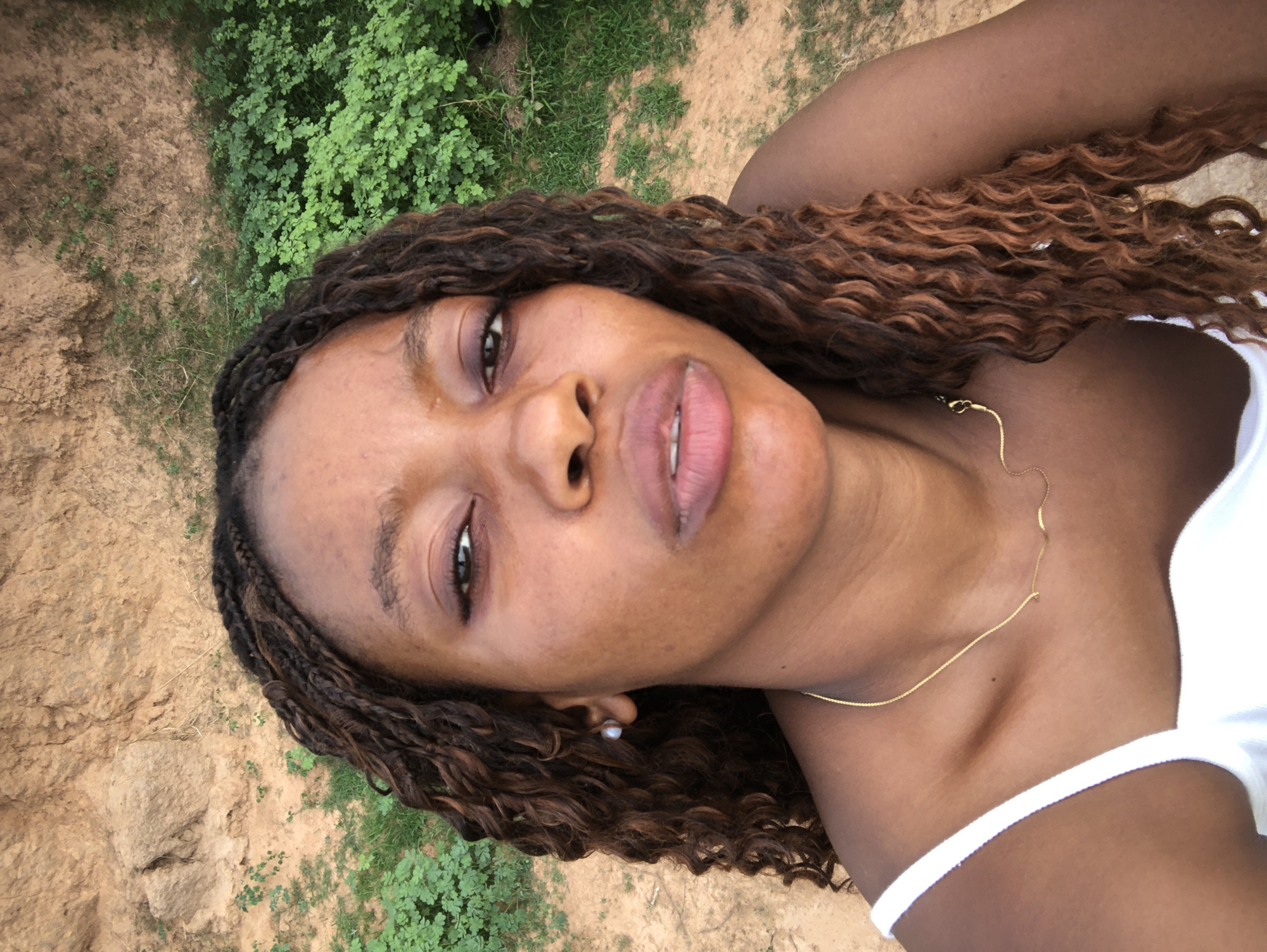 A medium close-up portrait shows a young Black woman. Her hair is styled in long, dark brown and auburn braids that cascade over her right shoulder. She wears a white top with a thin strap. A delicate gold necklace rests on her collarbone. Her skin is smooth with subtle blemishes. She has dark brown eyes, full lips, and a small pearl earring on her left ear. The background consists of dry, reddish-brown earth and patches of green vegetation. The location is potentially Kashere, Nigeria, though not definitively confirmed by the image alone. The image is taken from a slightly low angle, giving a view of the sky. The image appears to be a selfie.
