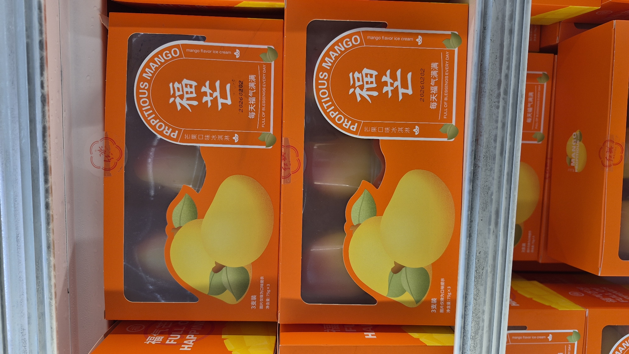 The image shows a close-up of several orange boxes of mango-flavored ice cream displayed in a retail setting, likely a grocery store freezer section. The boxes are stacked on shelves. Each box features an illustration of ripe mangoes and text in both English and Chinese. The English text includes "PROPITIOUS MANGO" and "FULL OF BLESSINGS EVERY DAY." The Chinese text includes "福芒" (Fú máng) which translates to propitious mango, and "每天福气满满" (Měitiān fúqì mǎnmǎn) which translates to full of blessings every day. Some boxes indicate "3支装" (3 zhī zhuāng), meaning a pack of 3, and a net weight of "75g×3". The setting appears to be indoors, with the cold and metallic sheen of freezer shelves visible. The image is taken from a slightly elevated angle, looking down at the products. There are no people visible in the image. The background is out of focus, but suggests the interior of a commercial establishment. Without further information or context provided in the image, the specific location within Rancho Cordova, United States of America cannot be determined.