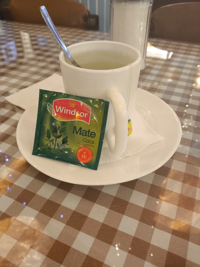 A white teacup with a silver spoon stands on a white saucer. A green Windsor Mate Coca tea bag packet leans against the cup's handle. The table beneath features a brown and white checkered pattern. In the background, a glass container possibly holds sugar, and glimpses of a blue and white striped wall and furniture suggest an indoor setting.