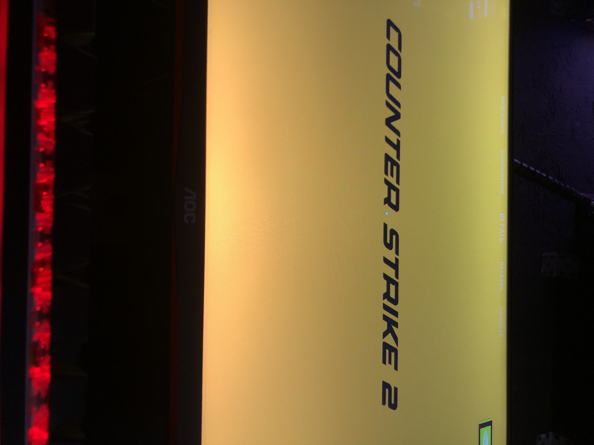 The image depicts a static computer screen displaying the main menu of the video game "COUNTER-STRIKE 2" in an indoor setting in Beryozovka, Russia. The monitor's screen shows a yellow-to-orange gradient background with the dark blue, stylized text "COUNTER-STRIKE 2" prominently centered. Above the title, five menu options are visible in white Cyrillic script: "ИНВЕНТАРЬ" (Inventory), "СНАРЯЖЕНИЕ" (Equipment), "ИГРАТЬ" (Play), "МАГАЗИН" (Shop), and "НОВОСТИ" (News). A small green rectangular icon is located in the upper right corner of the screen. The monitor itself is an AOC brand, with its logo visible on the bottom bezel. Below the monitor, a dark surface, consistent with a desk, shows illuminated red elements, indicative of a backlit gaming keyboard or peripheral. The area above the monitor is dark and out of focus, suggesting a dimly lit room. No individuals or active interactions are present in the scene.