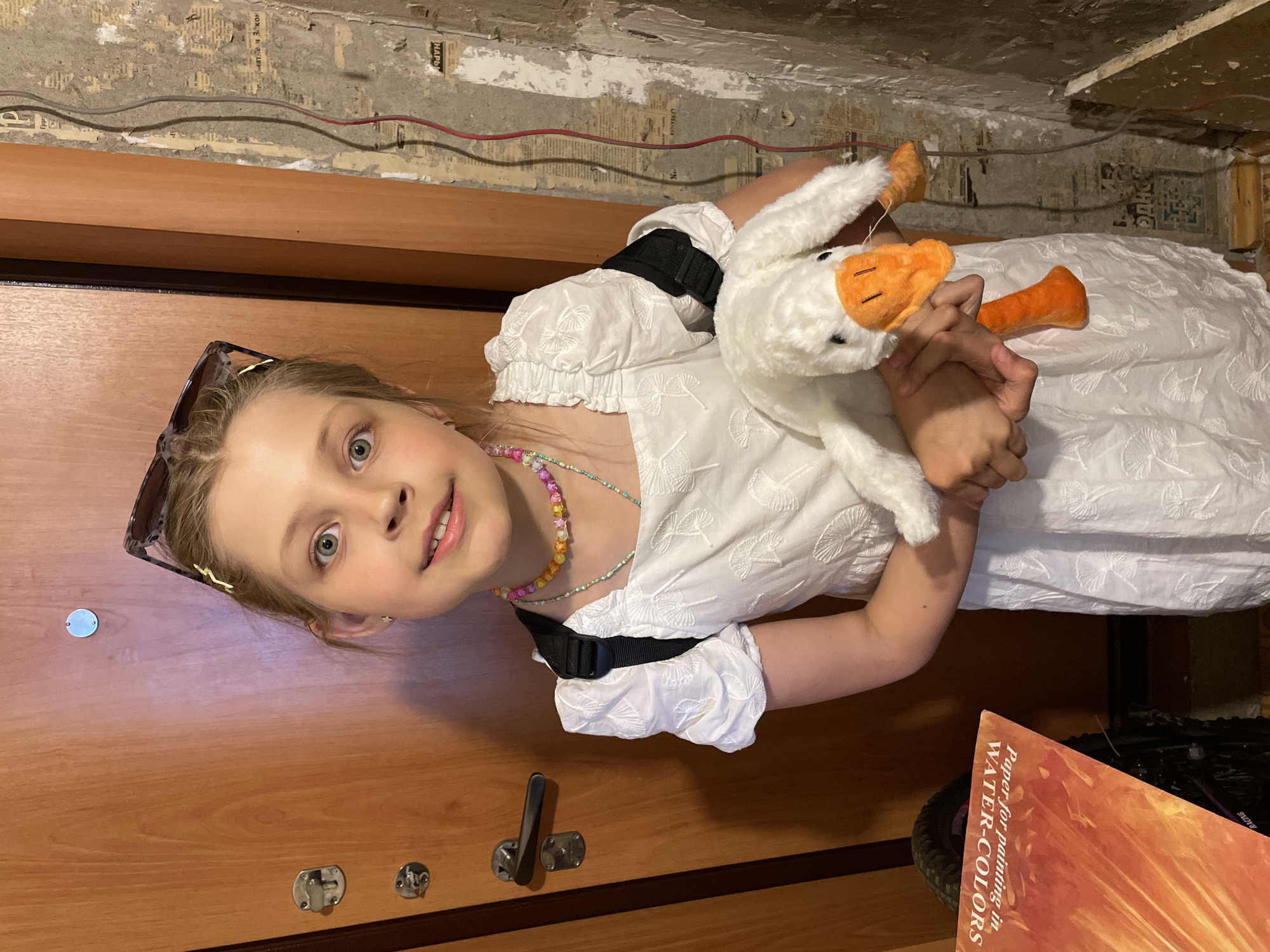 A young girl is holding a stuffed goose while standing inside a building. She is wearing a white dress and a colorful beaded necklace. Sunglasses are perched on her head. The background includes a wooden door, peeling wallpaper, and exposed wiring on the wall. The location is Izhevsk, Russia, given the presence of Cyrillic script on the wallpaper. A book titled "Paper for painting in WATER-COLORS" is visible in the lower right corner.