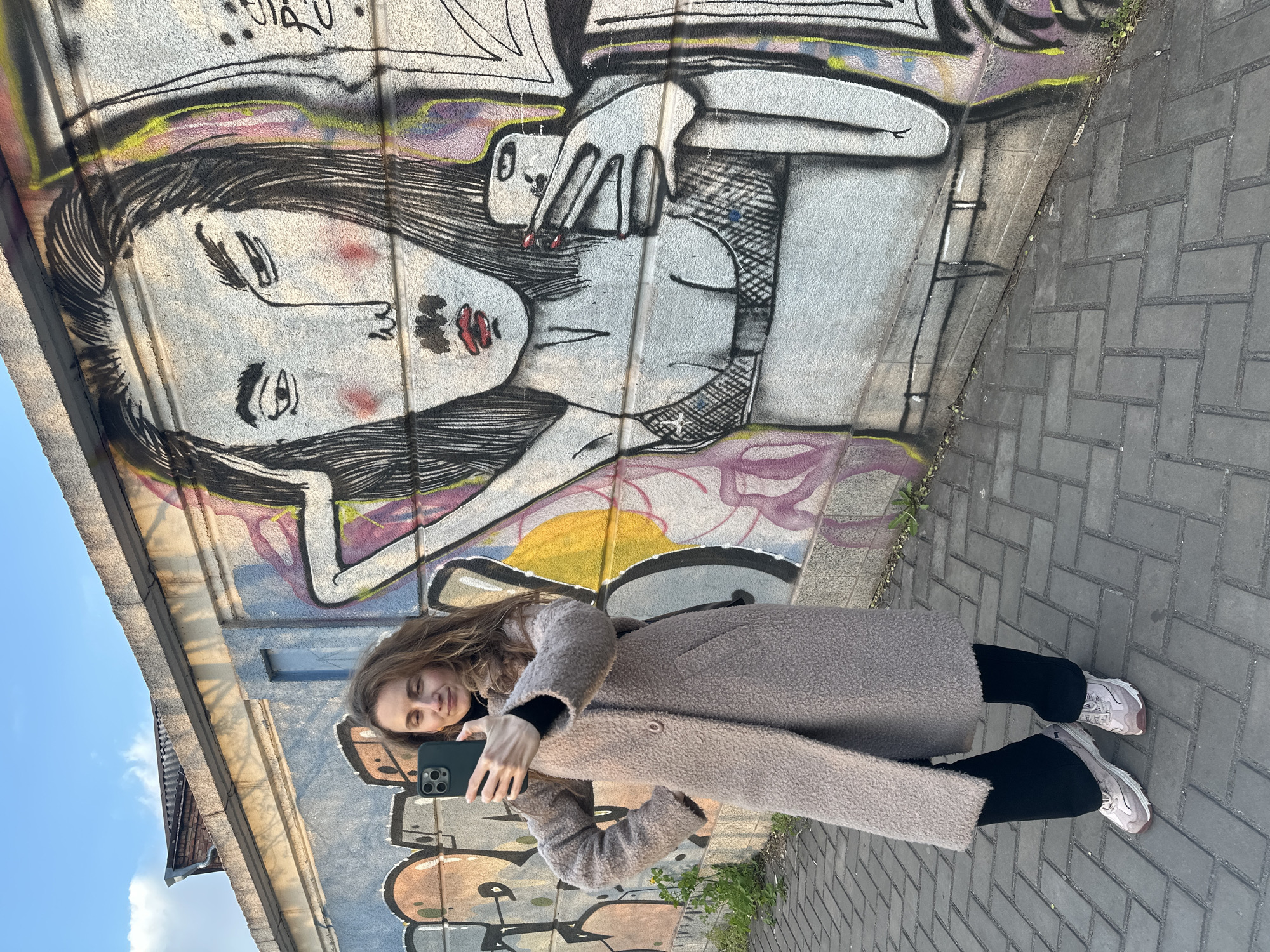 A young woman is depicted standing outdoors in Kryvyi Rih, Ukraine, on a paved grey brick sidewalk, against a vibrant, graffiti-covered concrete wall. She has long, wavy brown hair and is wearing a light brown or beige fluffy long coat over a black top, black trousers, and light-colored sneakers. She holds a dark smartphone, appearing to take a selfie or interact with the device, and has a relaxed, slight smile on her face.
The prominent graffiti behind her features a large, stylized illustration of a woman's face and upper body. This graffiti woman has long, dark hair, expressive eyes with long lashes, prominent red lips, and red blush on her cheeks. One hand is depicted with red painted nails, holding a thin object, possibly a cigarette. Part of her attire includes a fishnet-patterned top or bra. The graffiti incorporates splashes of yellow, purple, black, and pink colors.
The scene is set during the daytime under a clear blue sky, with visible shadows suggesting morning or afternoon. A building roof is partially visible in the upper left. No clearly legible text is discernible within the graffiti.