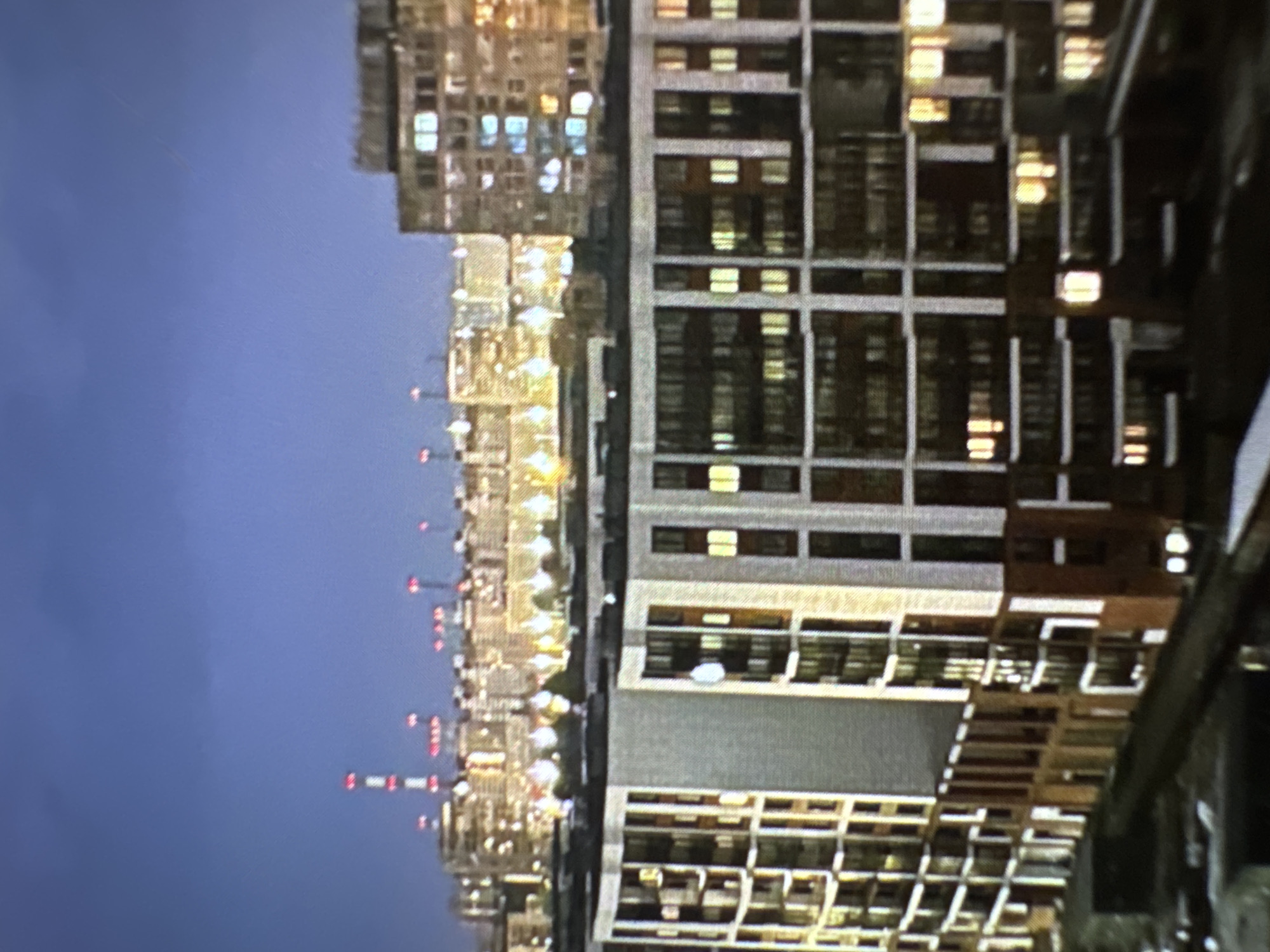 This image depicts a horizontal view of a city at what appears to be dusk or night, with the sky transitioning from a deep blue at the top to a darker shade towards the bottom. The scene is dominated by multiple tall buildings, some appearing as residential or office blocks with numerous illuminated windows, casting a warm glow. Other structures, possibly in the mid-ground, show a line of evenly spaced, smaller lights, and further in the distance, taller structures are topped with distinct red warning lights or beacons.
The buildings vary in architectural style; some have a grid-like facade with many small, square windows, while others appear more solid. There are no discernible people or specific activities taking place; the scene is a static cityscape. The sky is clear, with no clouds visible. The artificial lights from the buildings and the distant red lights stand out against the dark sky. No text is visible in the image. While the provided context indicates Chaikovsky, Russia, there are no specific landmarks within the image to confirm this location visually.