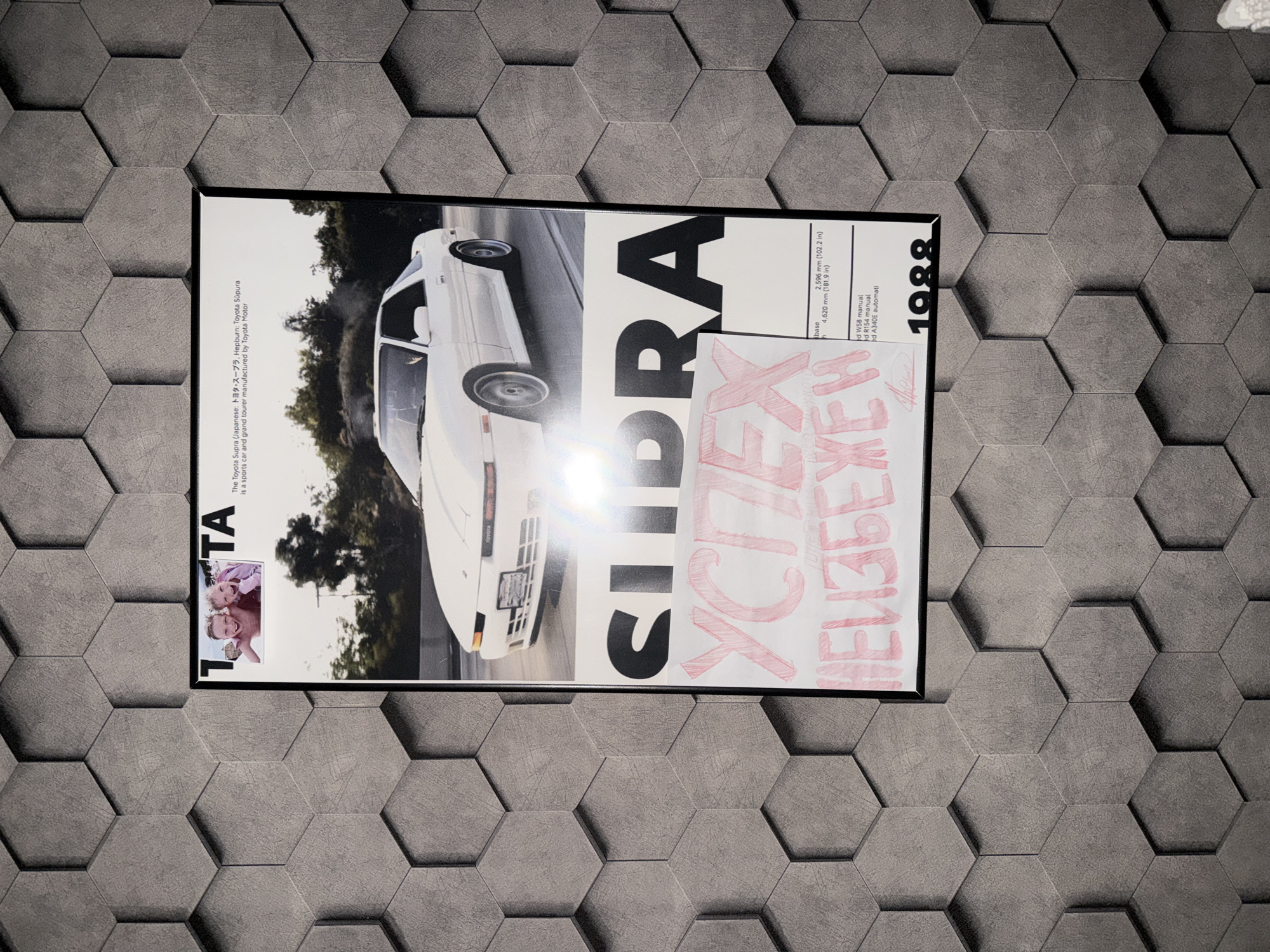 This is a photograph of a framed poster displayed on a textured wall composed of gray hexagonal tiles. The poster features an image of a white Toyota Supra car. Above the car, there are two children in a photograph. Text on the poster includes "TA", Japanese characters, and the phrase "The Toyota Supra (Japanese: トヨタ・スープラ Hepburn: Toyota Sūpura) is a sports car and grand tourer manufactured by Toyota Motor". Below the car image, the word "SUPRA" is prominently displayed in large black letters. Further down, there is writing in red marker that appears to be "УСПЕХ" and "НЕИЗБЕЖЕН". At the bottom right of the poster, the year "1988" is visible. There is a small inset image of a signature on the bottom left of the poster. The overall scene is indoors, with no discernible elements linking it to Nizhny Novgorod, Russia.