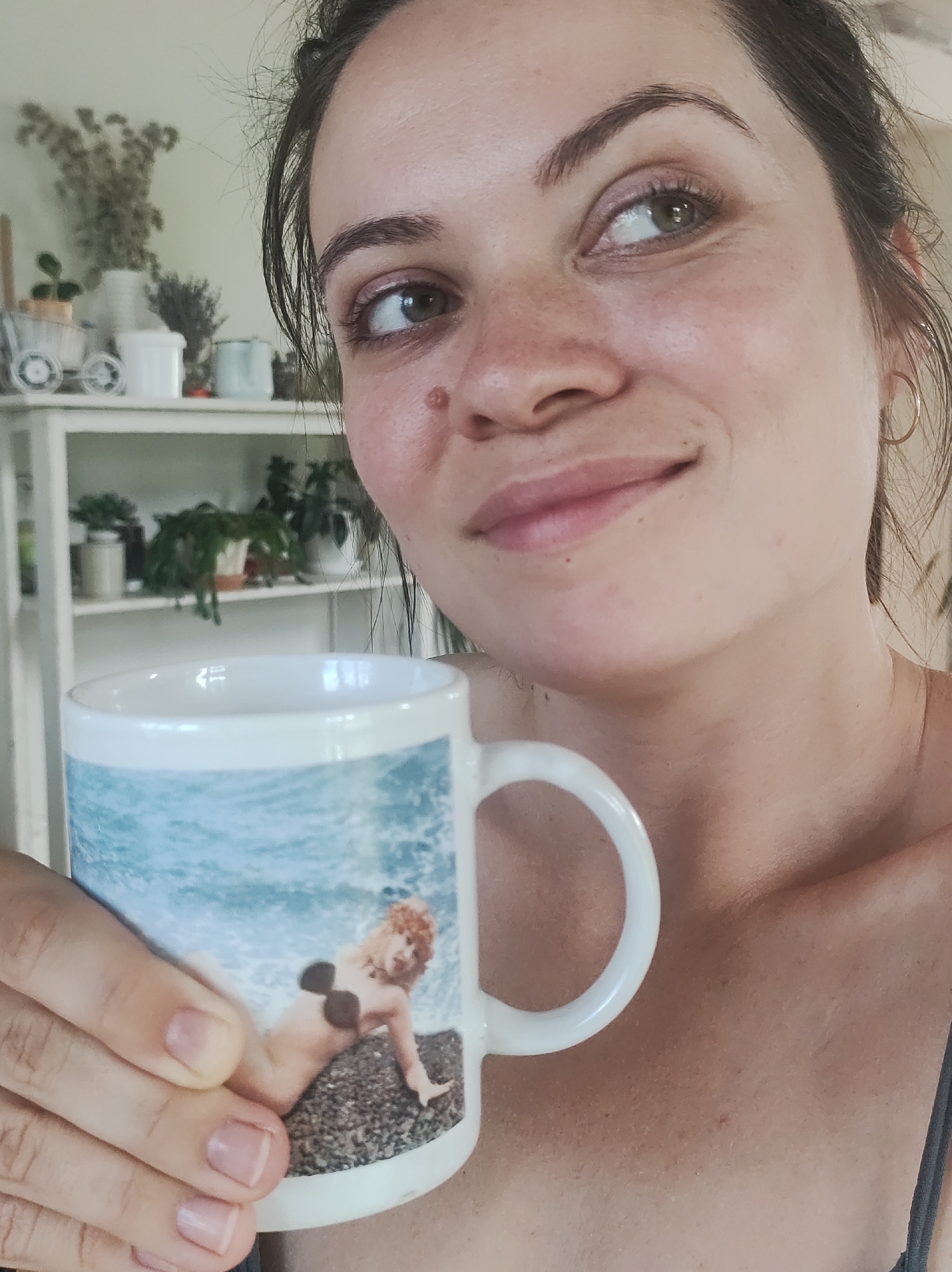 An adult female is depicted from the chest up, holding a white ceramic mug, in an indoor setting located in the city of Фоки, Russia. The female has dark hair, green-brown eyes, and fair skin with visible freckles and a raised dermal lesion on her right cheek. She wears a dark, thin-strapped top and a small gold hoop earring in her right ear. Her gaze is directed towards the upper right of the frame, accompanied by a subtle smile. Her unpainted fingernails are visible on the hands gripping the mug. The mug features a printed design depicting ocean waves above a rocky beach, where a cherubic figure with reddish-blonde hair and dark pasties reclines. The background consists of a light-colored wall and a white multi-shelf unit. The shelves display dried plant arrangements, a small white decorative wagon, various white containers, and multiple potted green plants.