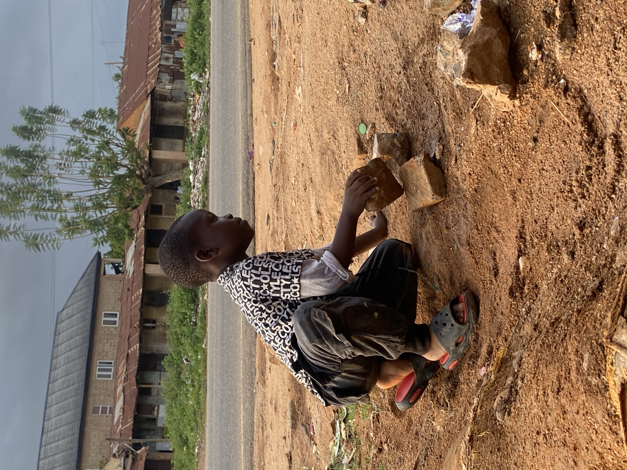 A young Black child wearing a black and white patterned shirt, black pants, and red and black sandals is squatting on a dirt surface near a road. The child is holding a brick in their hands and appears to be playing with it. The background shows a street with buildings, some in disrepair, and foliage. The sky is overcast. This image appears to have been taken in Vom, Nigeria.