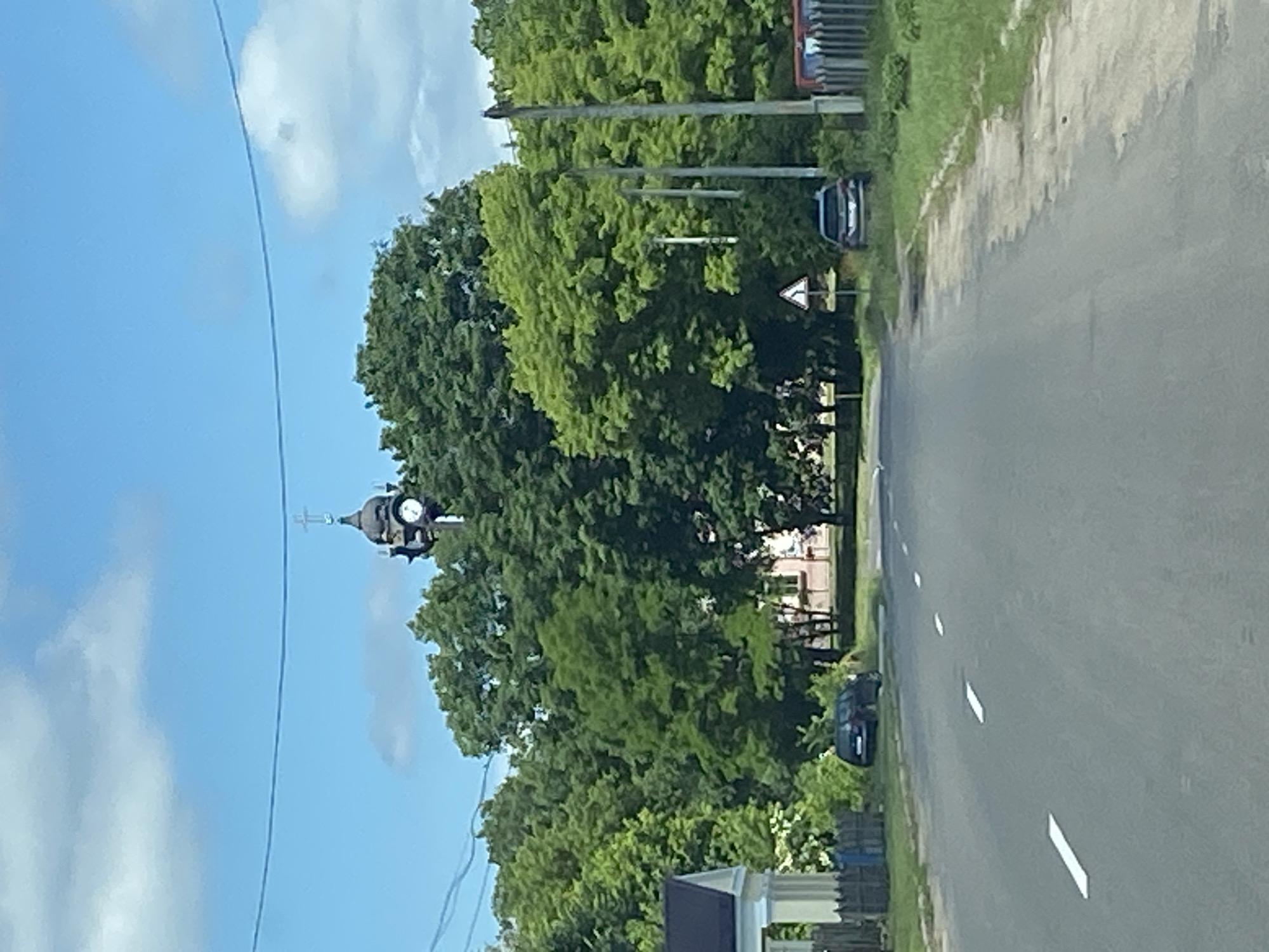 The image depicts a rural road in Воўчынскі сельскі Савет, Belarus, on a clear, cloudy day. The road is paved and has white dashed lines marking the lanes. On the left side of the road, a white building with a dark roof and columns is partially visible, with a wooden fence in front of it. Further down the road, a dark-colored car is parked on the side. On the right side, another dark-colored car is parked near a triangular road sign. A row of tall trees lines both sides of the road, with a church tower topped with a cross and a clock visible above the trees in the distance. Utility poles are also visible along the right side of the road. The overall scene conveys a sense of tranquility in a rural setting.