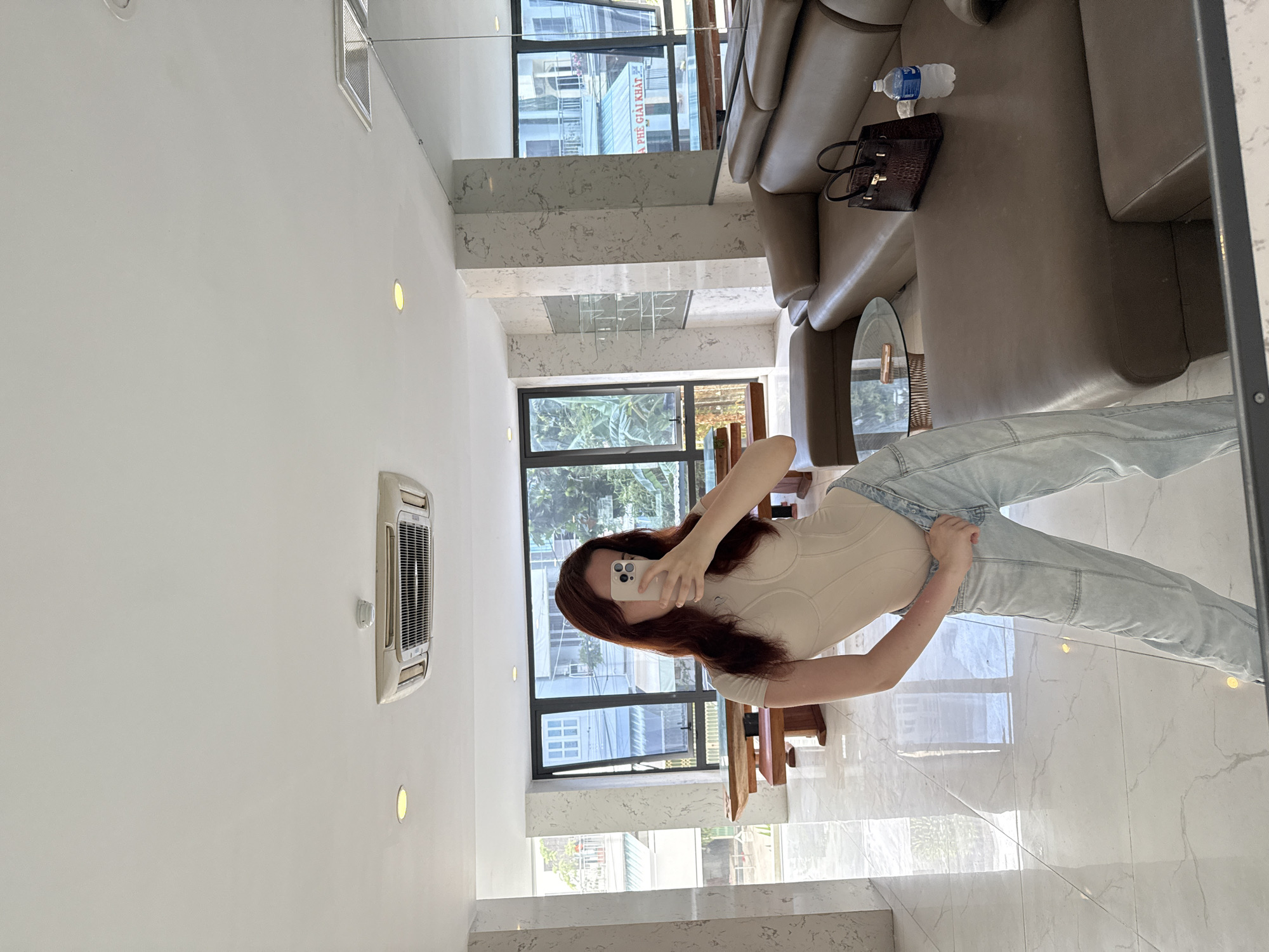 A woman is taking a mirror selfie in what appears to be a modernly designed interior. The location seems to be in Bac Nha Trang Ward, Vietnam, based on the reflection of buildings visible through the windows.
The woman is holding a smartphone in front of her face. She's wearing a light-colored top and light-wash denim jeans. Her dark hair is partially obscuring her face.
The interior includes a beige leather sofa with a dark brown handbag and a bottle of water placed on it. A small glass-topped table is beside the sofa. The walls are white and there is an air conditioner unit mounted on one. Large windows provide natural light and views of the city. The floor is covered with white marble tiles.