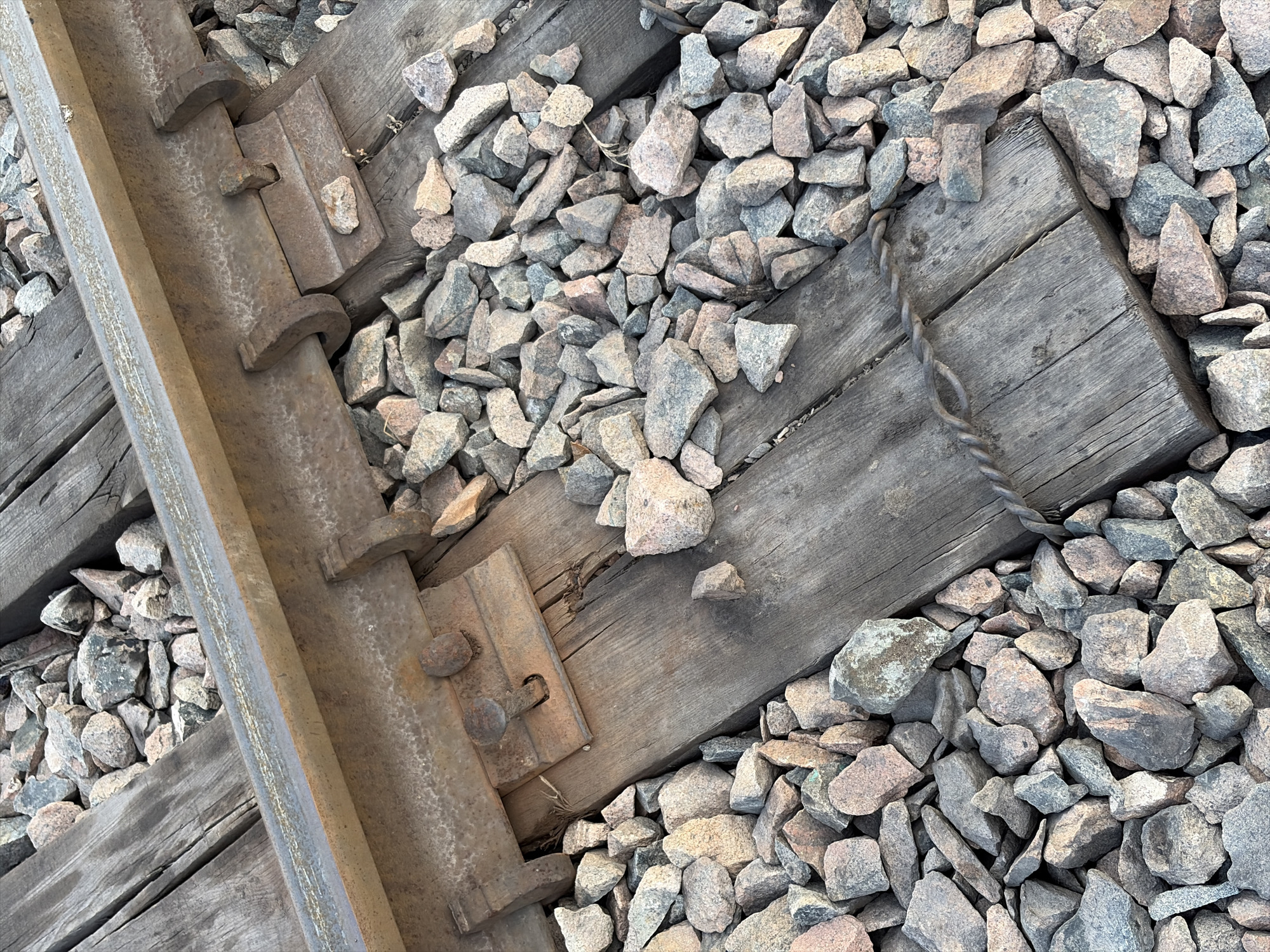 The image shows a close-up, overhead view of a section of railroad tracks. The primary subjects are the metal rails, wooden railway ties, and the gravel ballast that supports the tracks. The scene is outdoors, with the gravel composed of various shades of grey, pink, and tan rocks. The wooden ties appear weathered and worn, with visible grain and some splintering. The metal components, including the rail and fastening clips, are rusted and textured.
There is no visible activity or event taking place; the image captures a static, detailed view of the railway infrastructure. Notable details include the way the gravel fills the spaces between the ties and around the rails, suggesting a functional railway line. The lighting appears to be natural daylight, but there are no specific cues to determine the time of day or weather conditions. The location context of Kant, Kyrgyzstan, is provided but not visually evident in the image itself. There is no visible text within the image. The overall impression is one of rugged, industrial construction and the passage of time, conveyed through the textures and wear of the materials.