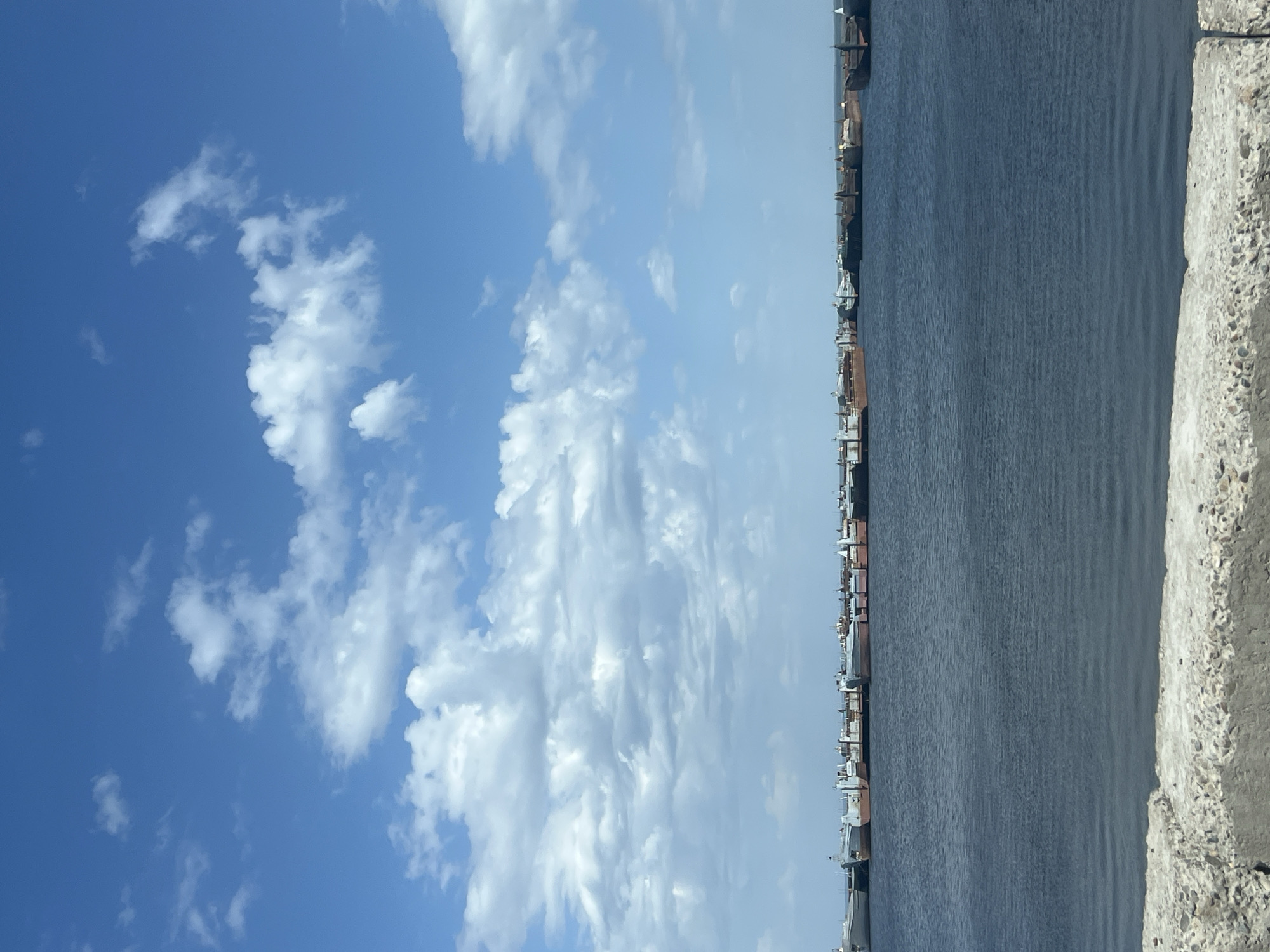The image depicts a view from an elevated position in the city of Chaikovsky, Russia. The scene is split vertically, with a bright blue sky and fluffy white clouds occupying the left half. The right half is dominated by a body of dark blue water, likely the Kama River, and features several docked ships and industrial buildings lining the shore in the distance. A concrete structure, likely part of a bridge or embankment, is visible in the foreground along the right edge of the image.