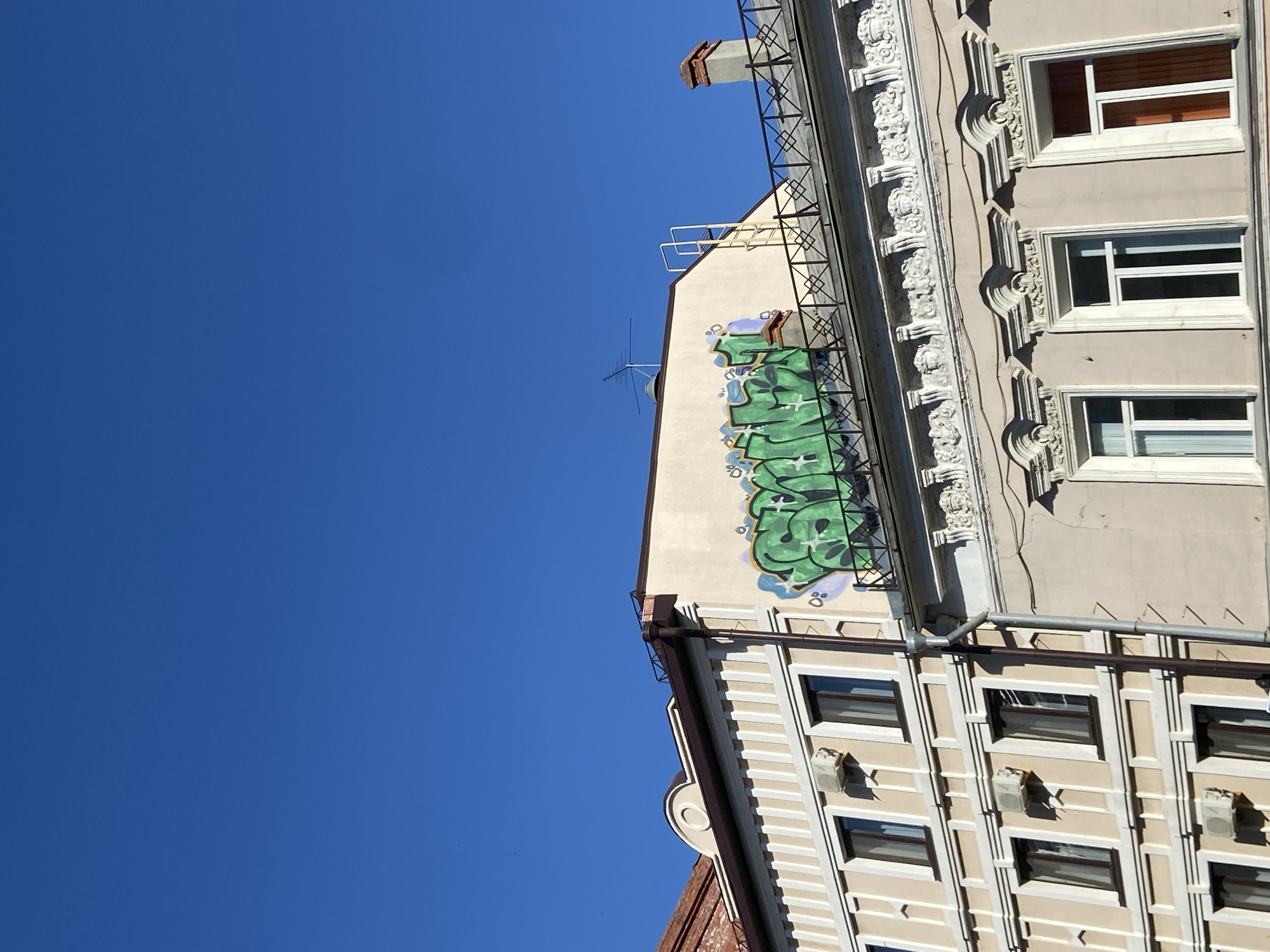 An upward-angled view shows portions of two adjacent buildings under a clear blue sky.
The building on the right, a light beige color, features multiple stories of windows with dark frames and light trim. Decorative molding is visible between the window rows. Rain gutters run vertically along the building.
The building on the left is a light tan color and has green and purple graffiti on the side visible. A metal fire escape ladder is mounted on the building. A television antenna extends from the top of the building.
The image captures a typical urban scene and is a generic street view. The exact location cannot be forensically determined without more evidence.