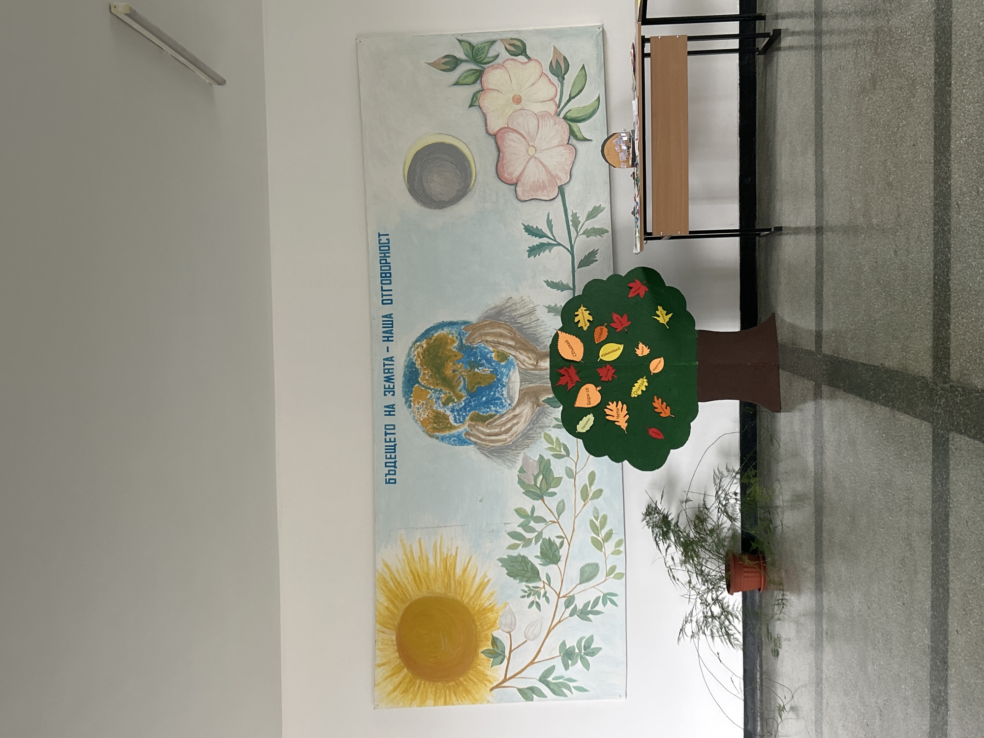 The image shows a wall in a building in Zlatograd, Bulgaria. On the wall is a large, vertically oriented painting or poster. The artwork depicts an image of the Earth being held in hands, with a sunflower at the bottom, a solar eclipse at the top, and pink flowers next to it. To the right of the painting is a coat rack and a cutout of a tree with fall leaves on it. To the right of the tree is a potted plant. The floor is tiled.