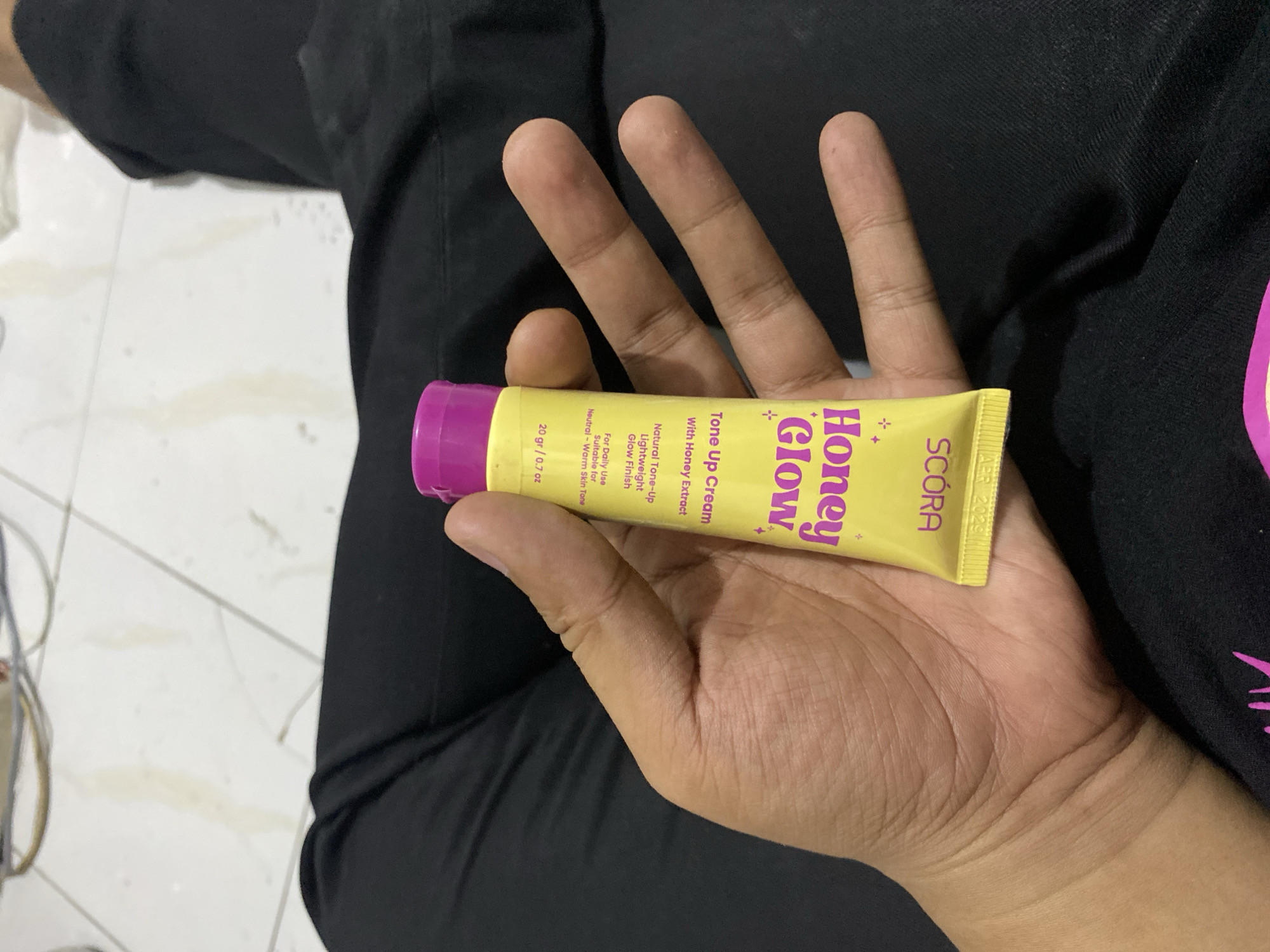A person's hand holds a small, yellow squeeze tube of "Honey Glow" tone-up cream. The tube has purple text and a purple cap. The person is wearing black clothing, and the background appears to be a tiled floor with some light-colored fabric. The tube is positioned diagonally across the palm of the hand, with the fingers wrapped around it. The image is taken from a close-up perspective, focusing on the hand and the product. The setting does not provide specific information about the city of Tebel, Indonesia.