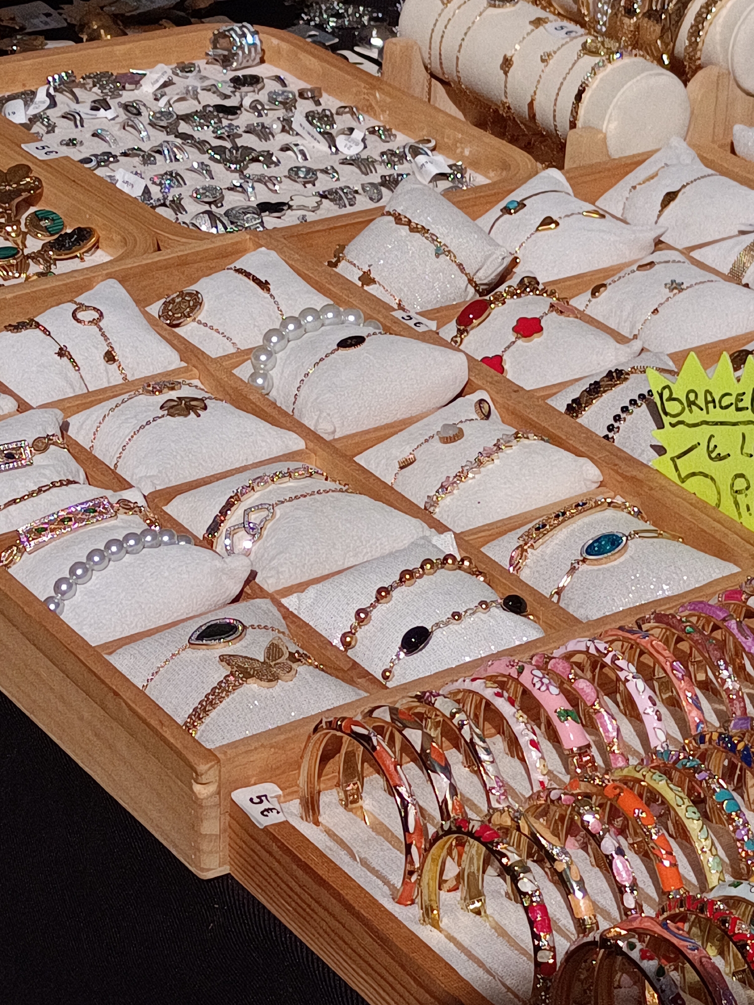 The image shows a stall at the market in Le Neubourg, France, displaying a variety of jewelry. The stall is made of wood and is divided into compartments, each showcasing different types of bracelets and rings. Some bracelets are adorned with pearls, while others feature colorful beads, gemstones, or intricate metalwork. There are also rings with decorative stones. A bright yellow price tag is visible, indicating "Bracelets 6€ 5€". In the foreground, a row of colorful bracelets with floral patterns are displayed. The overall scene suggests a bustling market environment.