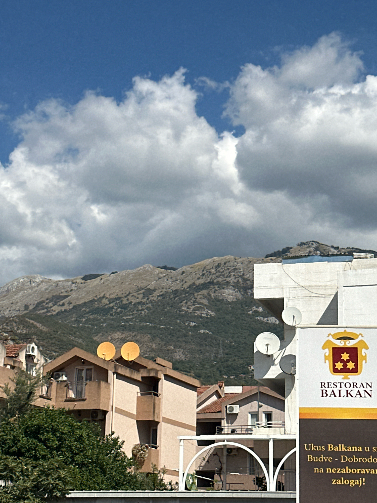 The image depicts a view of buildings and mountains under a cloudy sky in Budva, Montenegro. In the foreground, there are several buildings with apartment-like structures, some featuring balconies and satellite dishes. One building has a large advertisement for "RESTORAN BALKAN" with text in a foreign language. Behind these buildings, a large, rugged mountain range dominates the landscape, with patches of green vegetation on its slopes. The sky is filled with a mix of bright blue and large, fluffy white clouds, with some darker, more ominous clouds visible towards the mountains. The overall scene appears to be a typical urban and natural landscape of a coastal city.