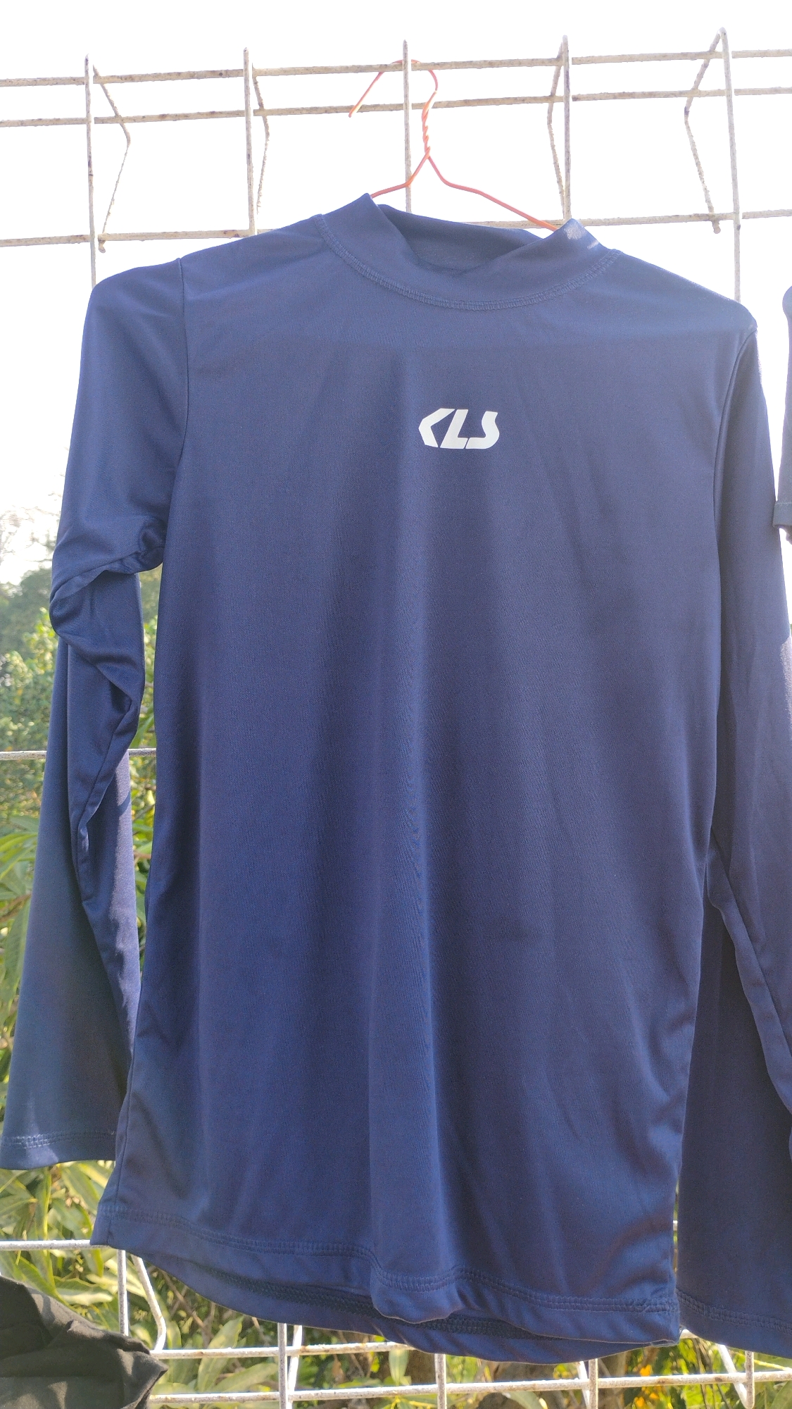 A navy blue long-sleeved shirt with a white logo is hanging on a clothesline outdoors. The shirt has a mock neck and is made of a lightweight, synthetic fabric. The logo, a stylized "CLS", is positioned on the left chest. The shirt is in focus, while the background, consisting of trees and a wire fence, is blurred. The scene is likely in Gumpang, Indonesia, suggested by the lush greenery and the bright, sunny atmosphere.