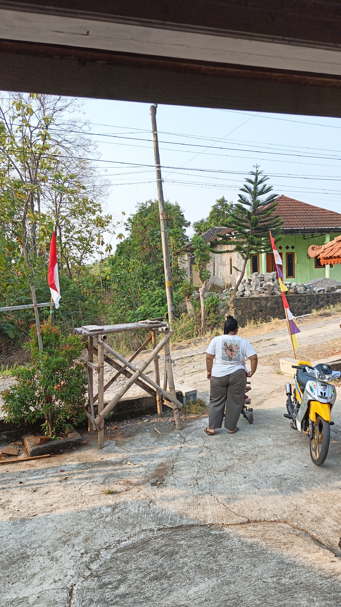 This image captures an outdoor scene in Sambi, Indonesia. A person, viewed from behind, is wearing a white t-shirt with a graphic design and olive-colored pants. They are standing next to a small, yellow and silver motorcycle. In the background, there are several houses, one of which is painted green. A tall pine tree stands in front of the green house. The ground is a mix of concrete and dirt. A wooden structure, possibly a scaffolding or platform, is visible to the left of the person. A red and white Indonesian flag is planted on a bamboo pole to the left of the wooden structure. Another flag, with multiple colors including red, white, and purple, is visible near the green house. The environment appears to be rural or semi-rural, with trees and foliage surrounding the structures. The lighting suggests it is daytime, possibly late afternoon, with long shadows cast on the ground.