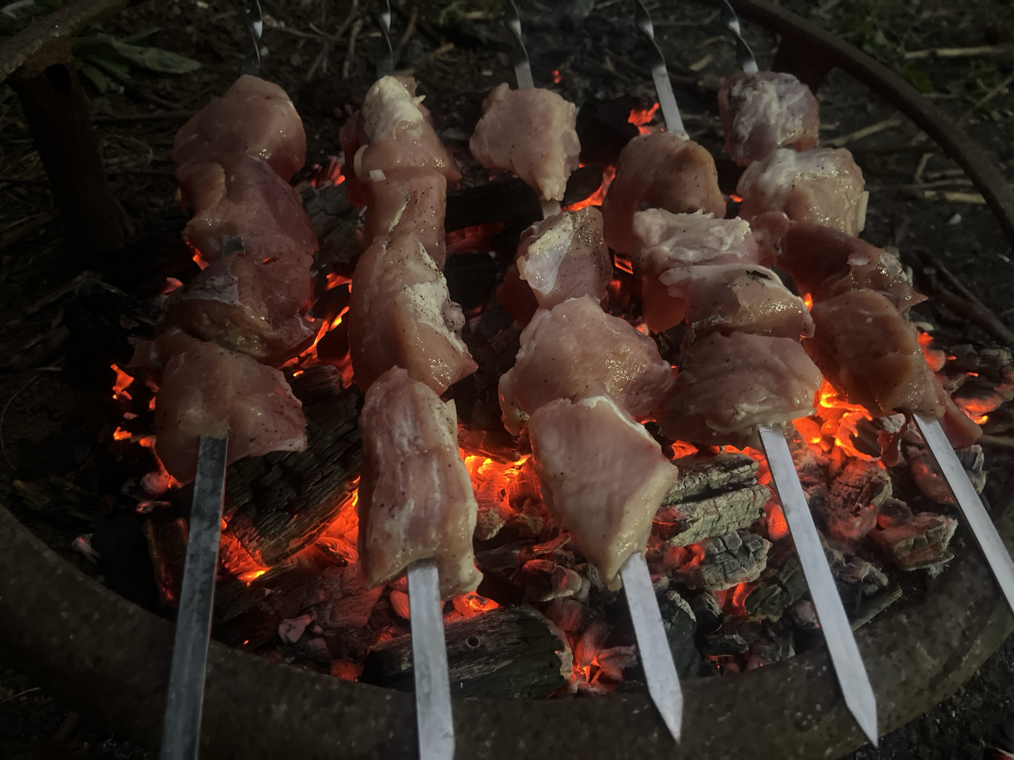 Four metal skewers laden with raw, cubed meat are positioned above a bed of glowing red coals. The meat, appearing to be pork or chicken, is seasoned with visible specks of black pepper and salt. The skewers are arranged vertically within a circular fire pit constructed from dark, rough material. The background is dark and out of focus, with hints of dry grass and foliage suggesting an outdoor, possibly rural, setting. The lighting is dim, with the intense glow of the coals illuminating the meat and creating strong shadows.