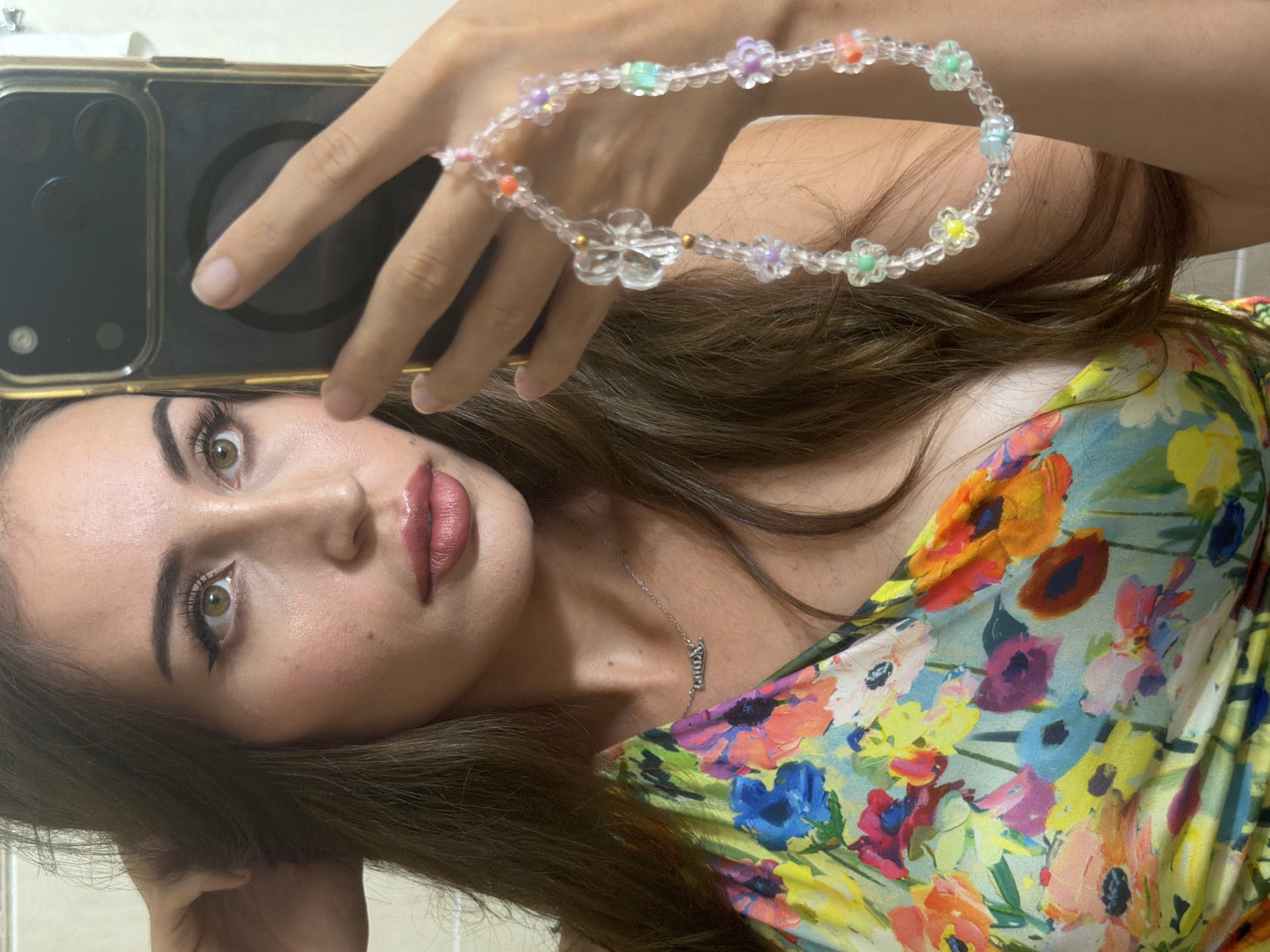 A young woman with long, dark brown hair and light brown eyes is taking a selfie in a brightly lit indoor setting. She is wearing a colorful floral print dress and a silver necklace with a pendant. Her right hand is holding a smartphone with a gold case, and her left hand is resting on her head. A beaded bracelet with flower charms is visible on her wrist. The background is blurred, but appears to be a tiled wall, possibly in a bathroom. There is no indication of the location in Tersefanou, Cyprus.