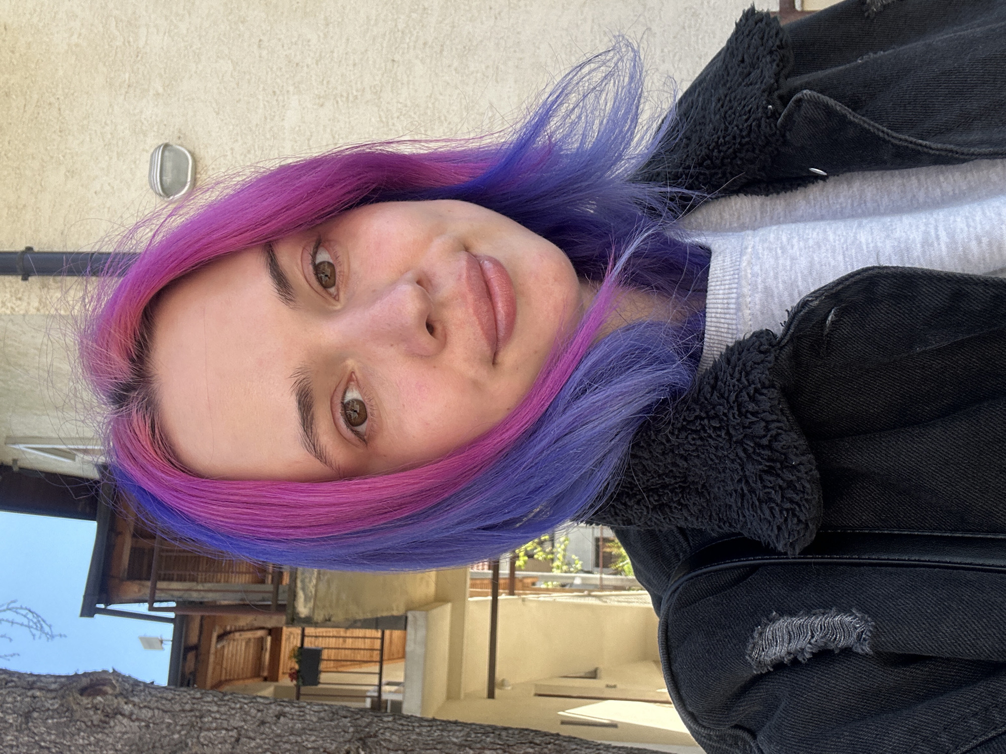 A young woman is centered in the frame, smiling and looking slightly downward. She has fair skin, light brown eyes, and her hair is dyed with a vibrant combination of purple and pink hues. She's wearing a grey sweatshirt and a black denim jacket with a dark, fuzzy lining. A patch of distressed denim is visible on the jacket near her shoulder.
The background suggests an outdoor urban setting. A light-colored building with a round outdoor light fixture is partially visible behind the woman. The bottom of the frame reveals a tree trunk, and further in the background, there's a glimpse of residential buildings and sky. The image appears to be taken in Zagreb, Croatia.