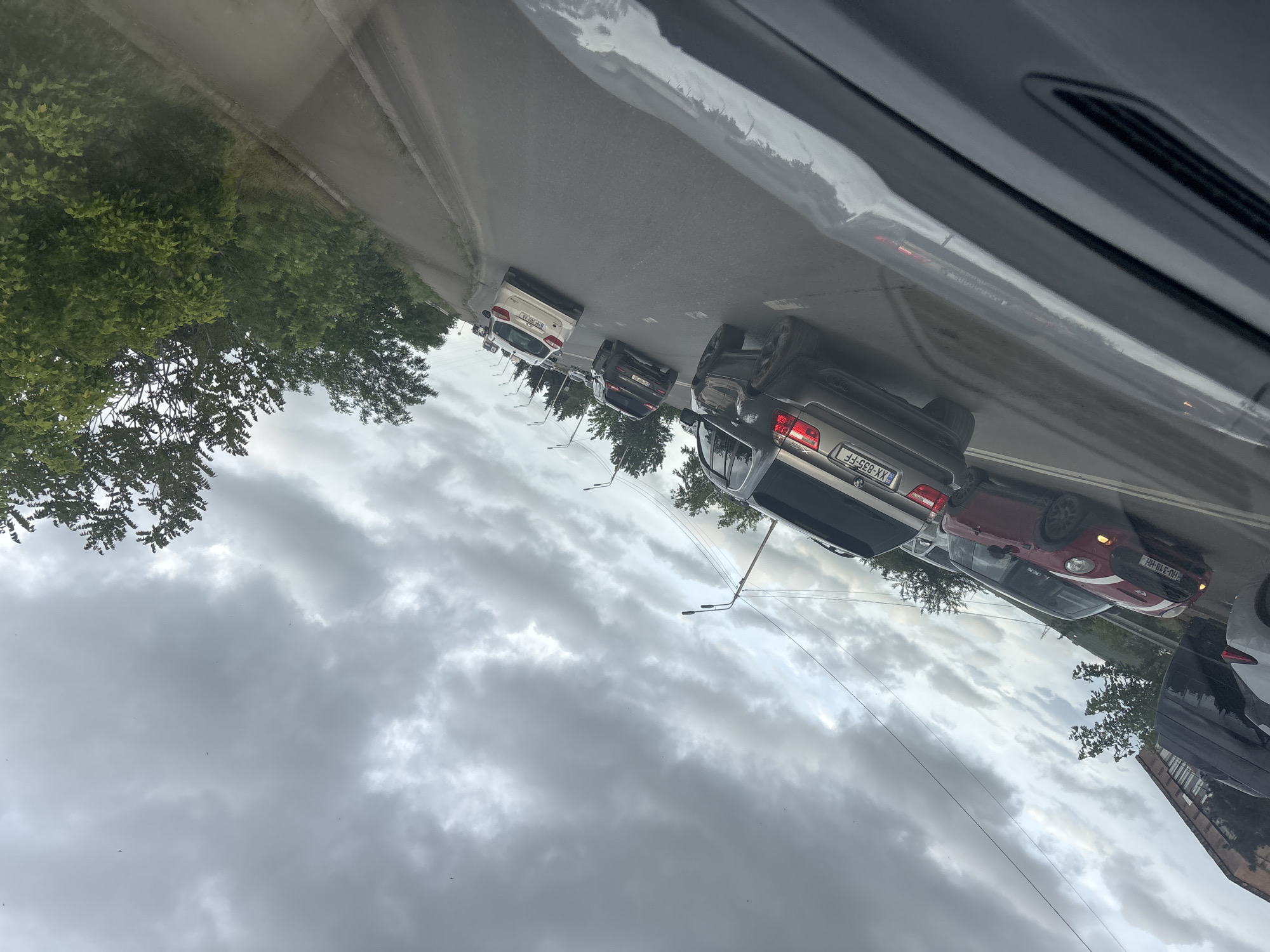 The image depicts a street scene in Tbilisi, Georgia, seen through the interior of a car. There is a line of vehicles on the road, including a gray Audi with license plate XX-835-FF, a red car with license plate HU-310-HH, a dark SUV, and a white car. The sky is overcast with cloudy weather. Trees are visible along the roadside. Streetlights line the road.