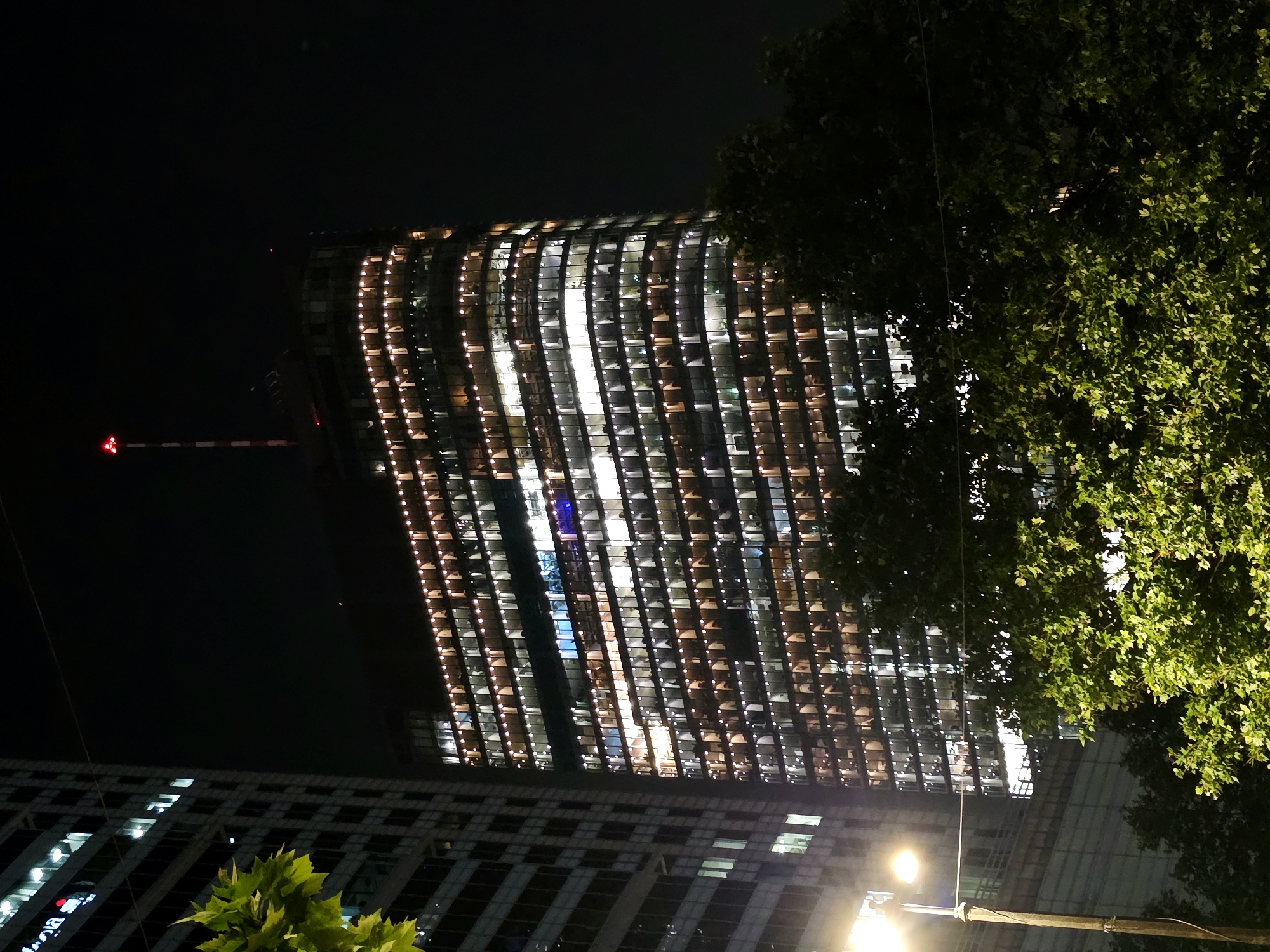 An exterior, low-angle, night-time shot shows a modern, illuminated skyscraper in Warsaw, Poland. The building is tall and rectangular, with a curved facade that is brightly lit by numerous interior lights visible through its windows. The lights are primarily a warm, yellowish-orange color, outlining the floors and the building's structure. At the very top of the skyscraper, a red blinking light is visible on a thin, vertical antenna or spire. To the left of the main skyscraper, another, taller, more angular building with fewer visible interior lights can be seen. In the foreground, lush green trees partially obscure the lower levels of the buildings. A street lamp with a bright, yellow light is visible on the left side of the frame, illuminating the foreground foliage. The scene is set against a dark night sky.