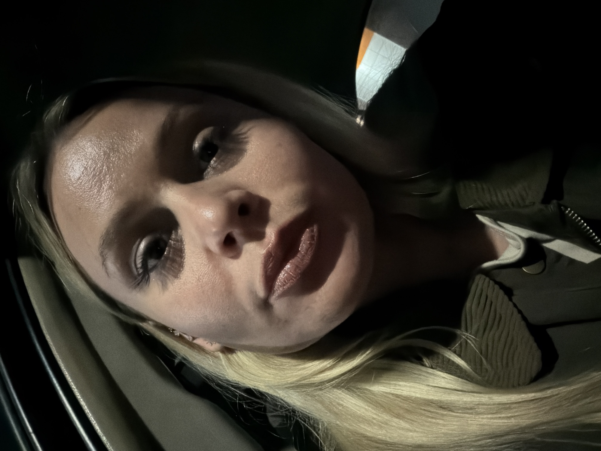 A close-up selfie depicts a fair-skinned woman with blonde hair, positioned at an oblique angle. Her hair is long and straight. Her facial features include prominent eyelashes, a defined nose, and full lips with visible lip gloss. She is wearing what appears to be an olive green jacket. The background is dark, suggesting a nighttime or low-light environment, and includes the interior of a vehicle, as evidenced by the car door frame visible on the left. There is no indication of the image being taken in Sarapul, Russia.