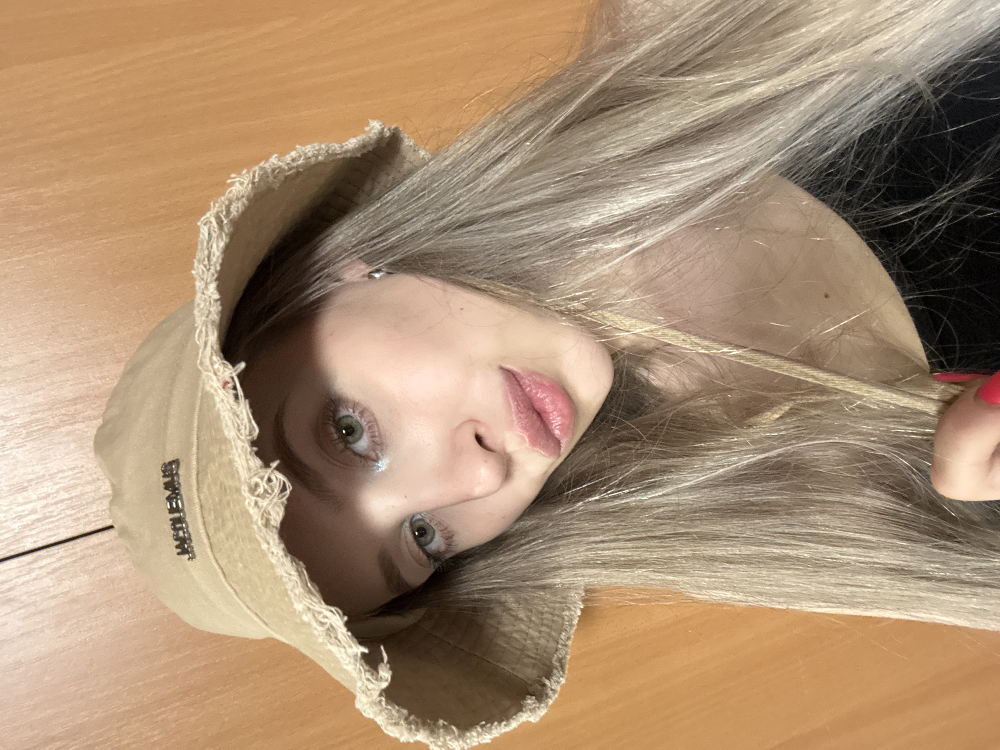 The image shows a close-up, top-down view of a young woman wearing a light-colored bucket hat with frayed edges. Her long, blonde hair spills over the hat and across her face. She is looking directly at the camera with wide, green eyes and her lips are slightly parted, revealing a glossy pink lipstick. The hat has a textured band with the word "JACQUEMUS" embossed in silver lettering. The background appears to be a light brown, wooden surface, possibly a desk or table, suggesting an indoor setting. The lighting is soft and even, casting no harsh shadows, and there are no clear indicators of the time of day or weather. The overall mood is casual and direct, with the subject making eye contact. The image was taken in Krasnoyarsk, Russia.