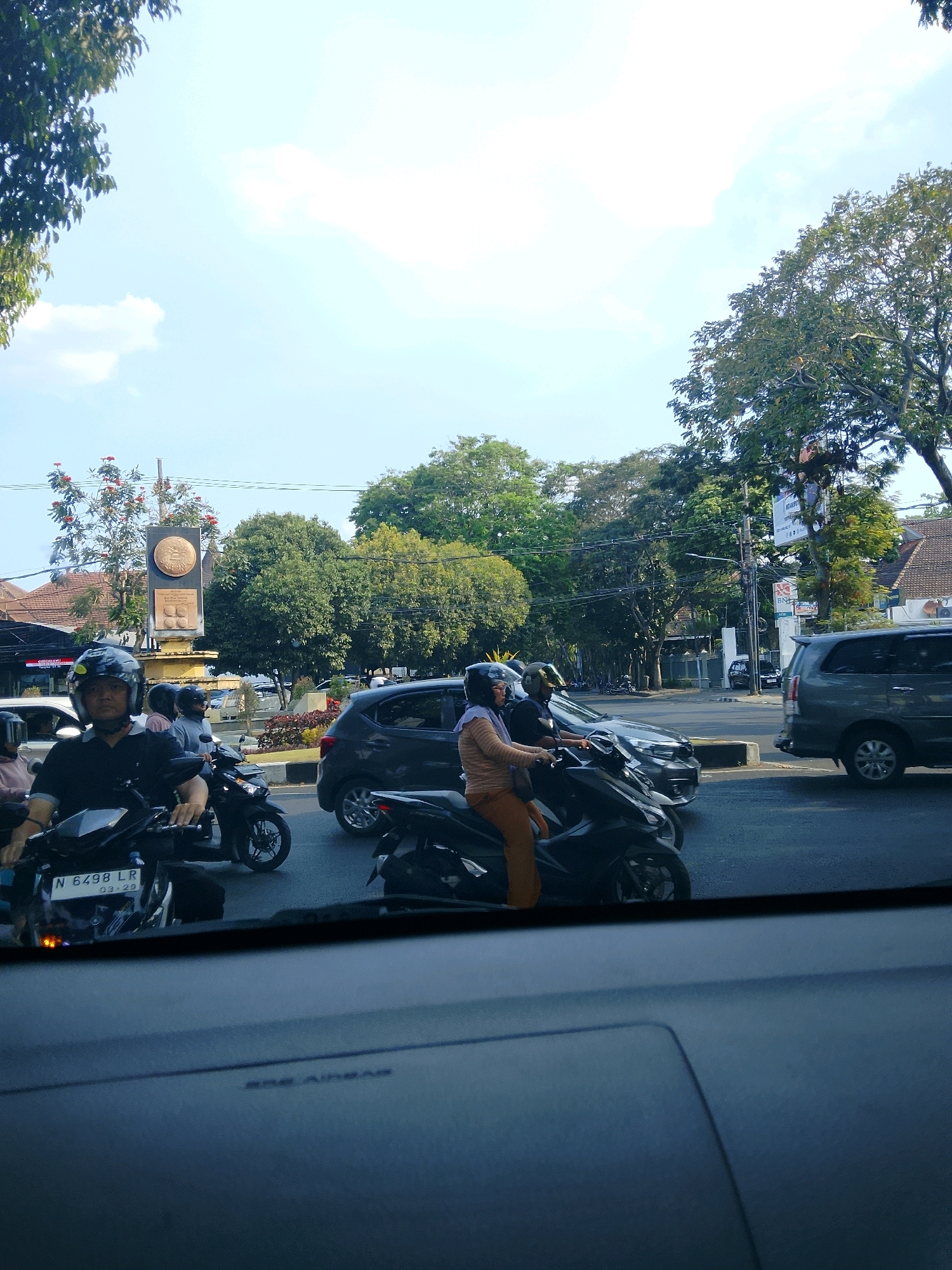 The image captures a street scene in Malang, Indonesia, likely during daylight hours, as indicated by the bright sky and shadows. The foreground shows the interior of a vehicle, with a dark blue dashboard in focus. Beyond the windshield, a traffic jam is visible, primarily composed of motorcycles and cars.
Several individuals are on motorcycles, all wearing helmets for safety. In the center of the frame, a man in a black shirt rides a motorcycle, with a license plate reading "N 6498 LR" and "03.23." To his right, a woman in an orange outfit is riding a scooter. Further back, other vehicles and motorcyclists are present.
The environment includes trees lining the street, providing a green canopy. A prominent monument or sculpture, likely a landmark in Malang, stands to the left of the road. It features a circular emblem at the top, with text below it. Buildings and shops are also visible in the background. The overall scene depicts a typical urban traffic situation in Indonesia.