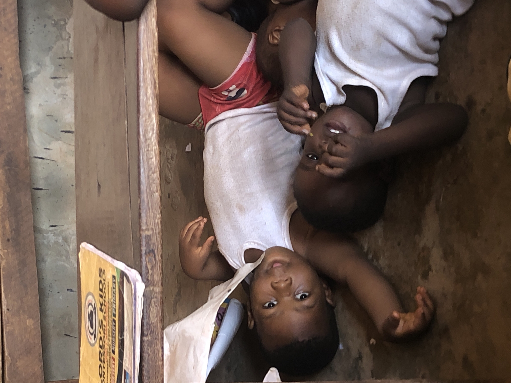 Two dark-skinned children wearing white tank tops and sitting in a small, enclosed area in Ukwu-Abwa, Nigeria, are visible. One child is wearing red shorts. A book titled "Lordis Kids Montessori Schools" is on the left. The area appears to be a corner or small room constructed of wood.